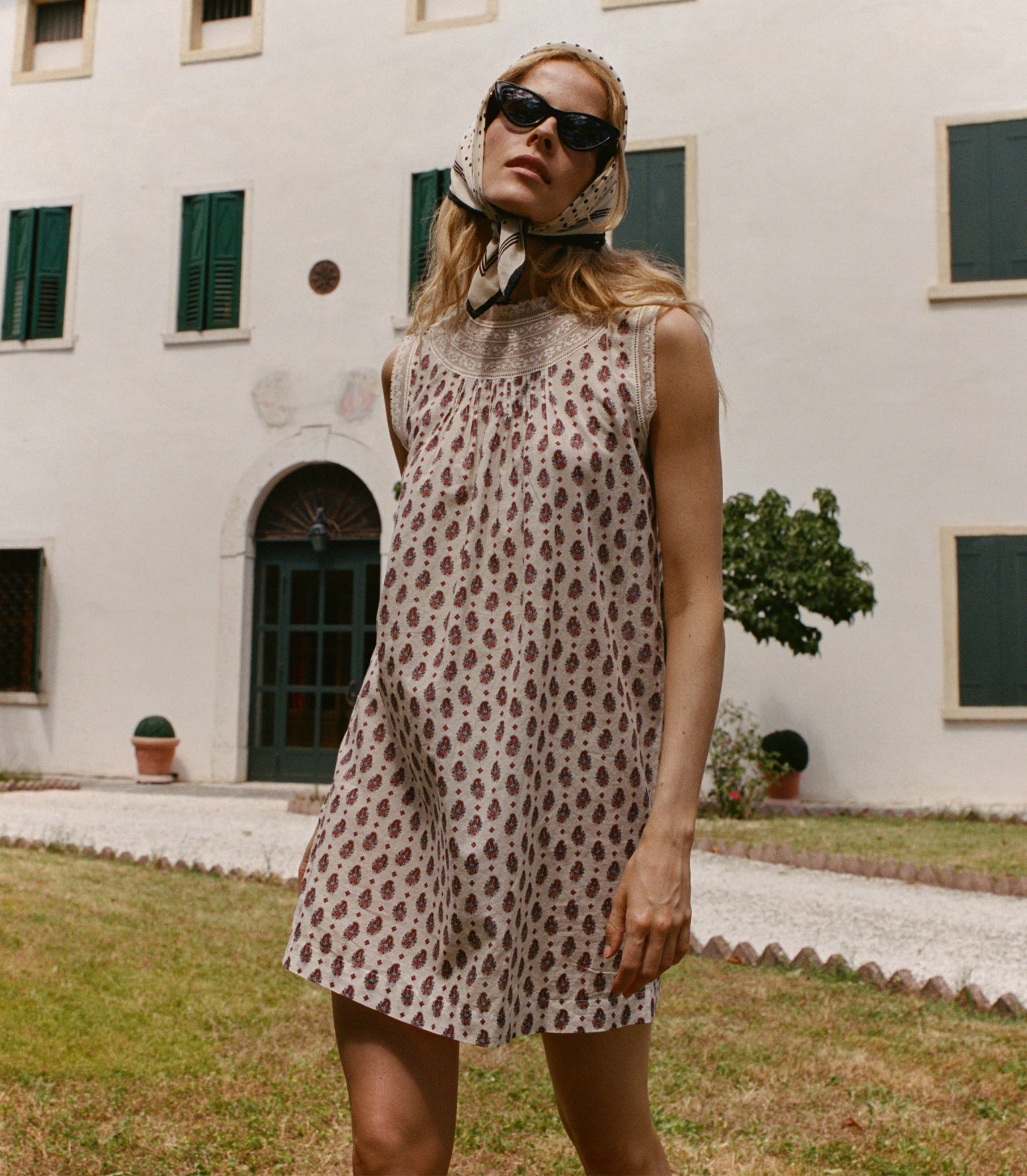Woman in a floral dress standing in front of a white building with green shutters.