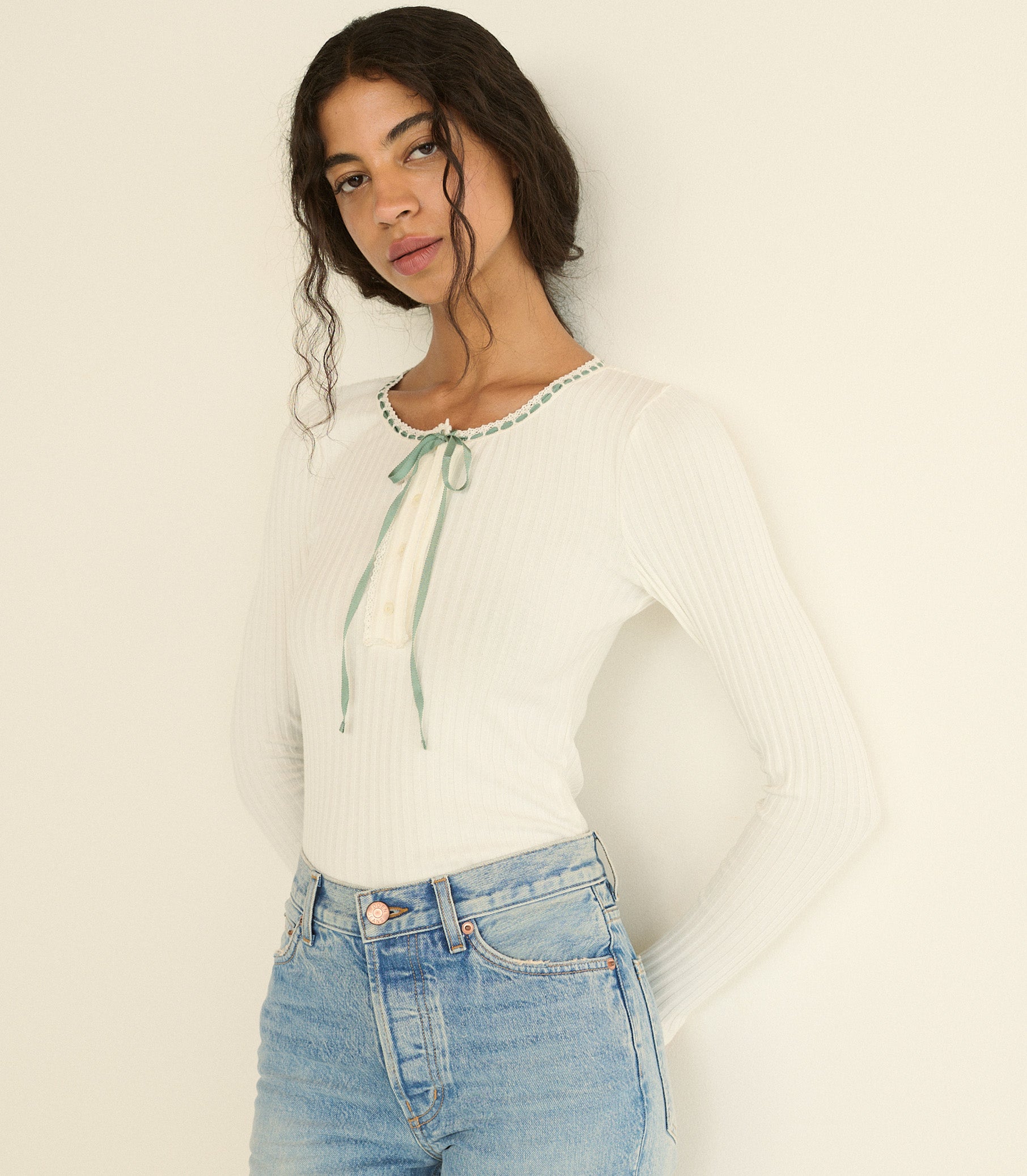 A woman wearing a white ribbed long-sleeve top with a mint blue tie detail at the neckline, paired with light blue jeans, against a plain off-white background. 1
