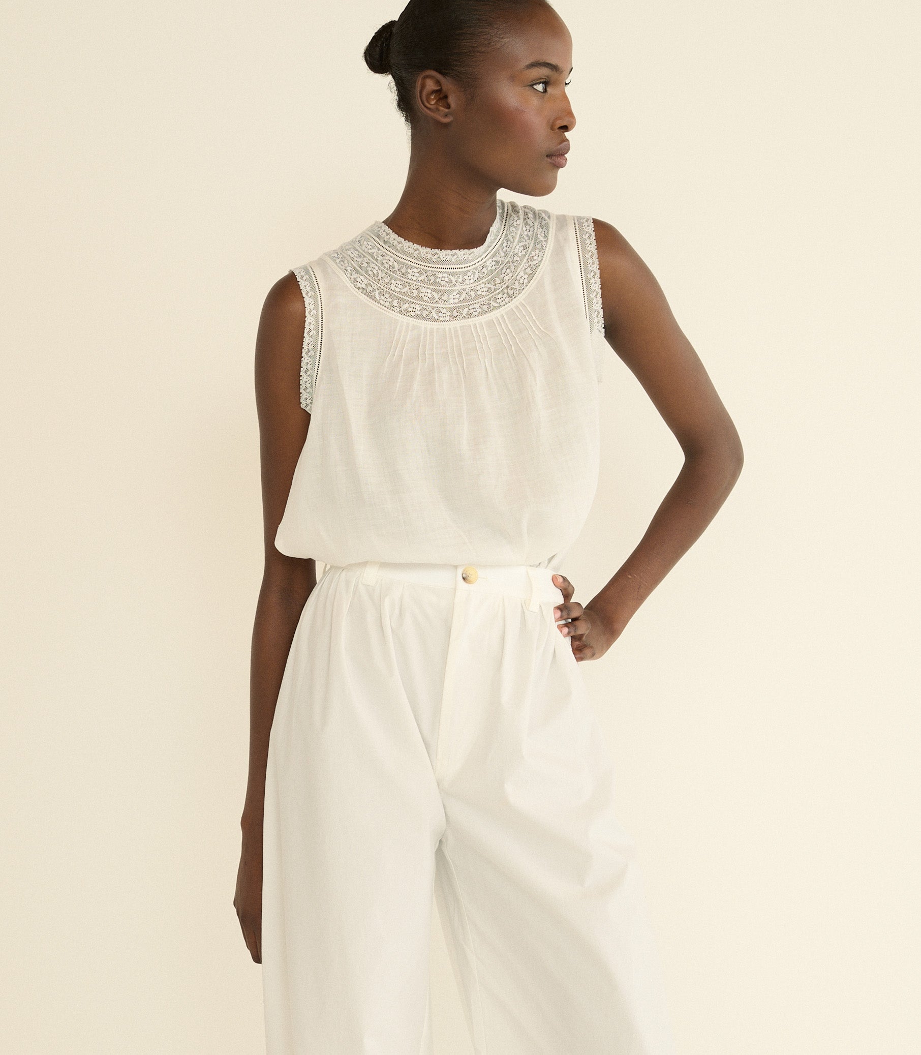 A model wearing a white sleeveless top with a detailed embroidered yoke and matching white wide-leg trousers, photographed against a solid light beige background. 1

