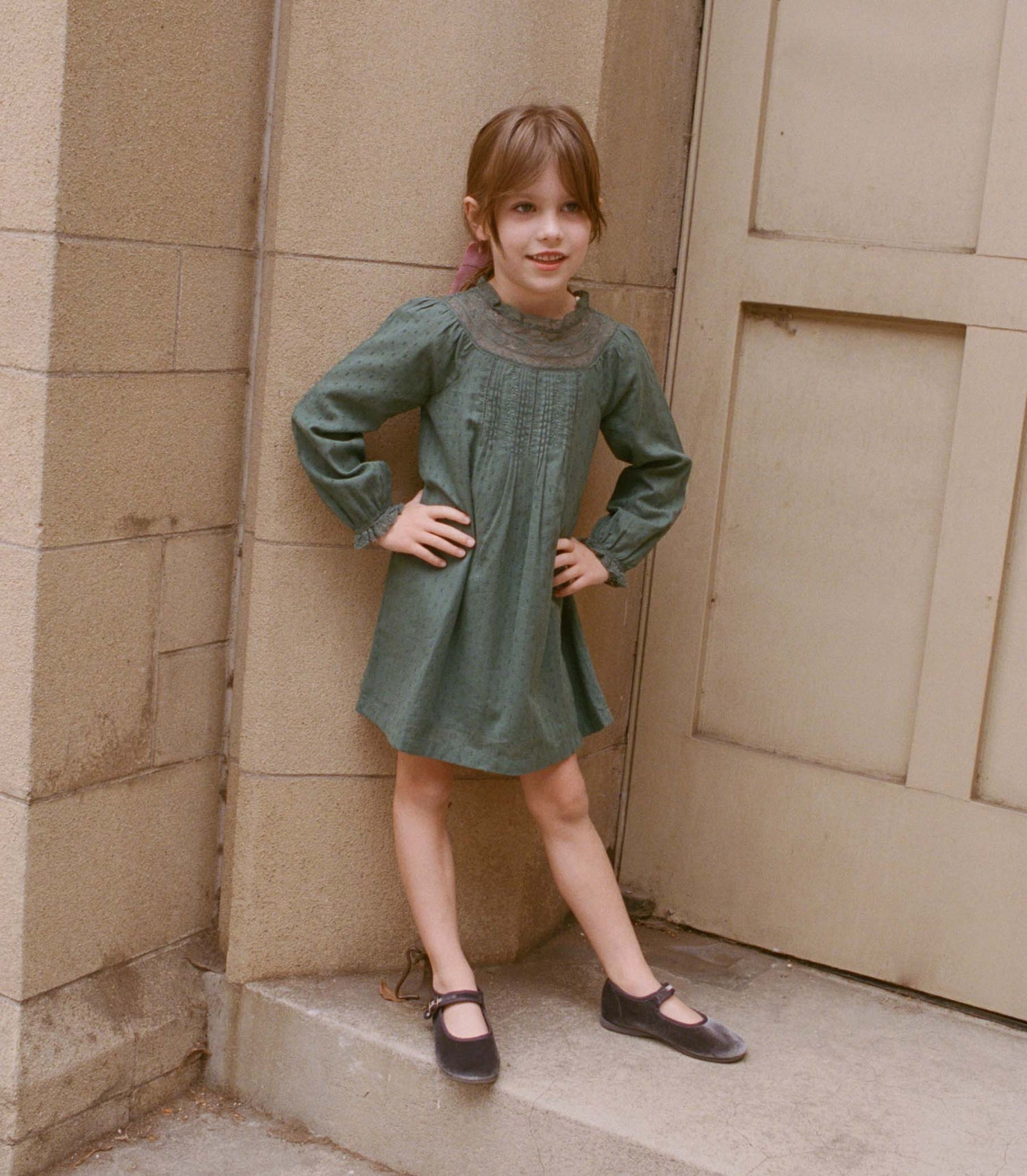 Young girl in a green dress standing against a beige wall.