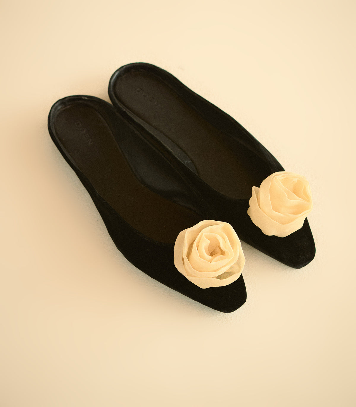 A pair of black pointed-toe flats with cream fabric rose embellishments on the outer side, displayed on a solid cream background. 1
