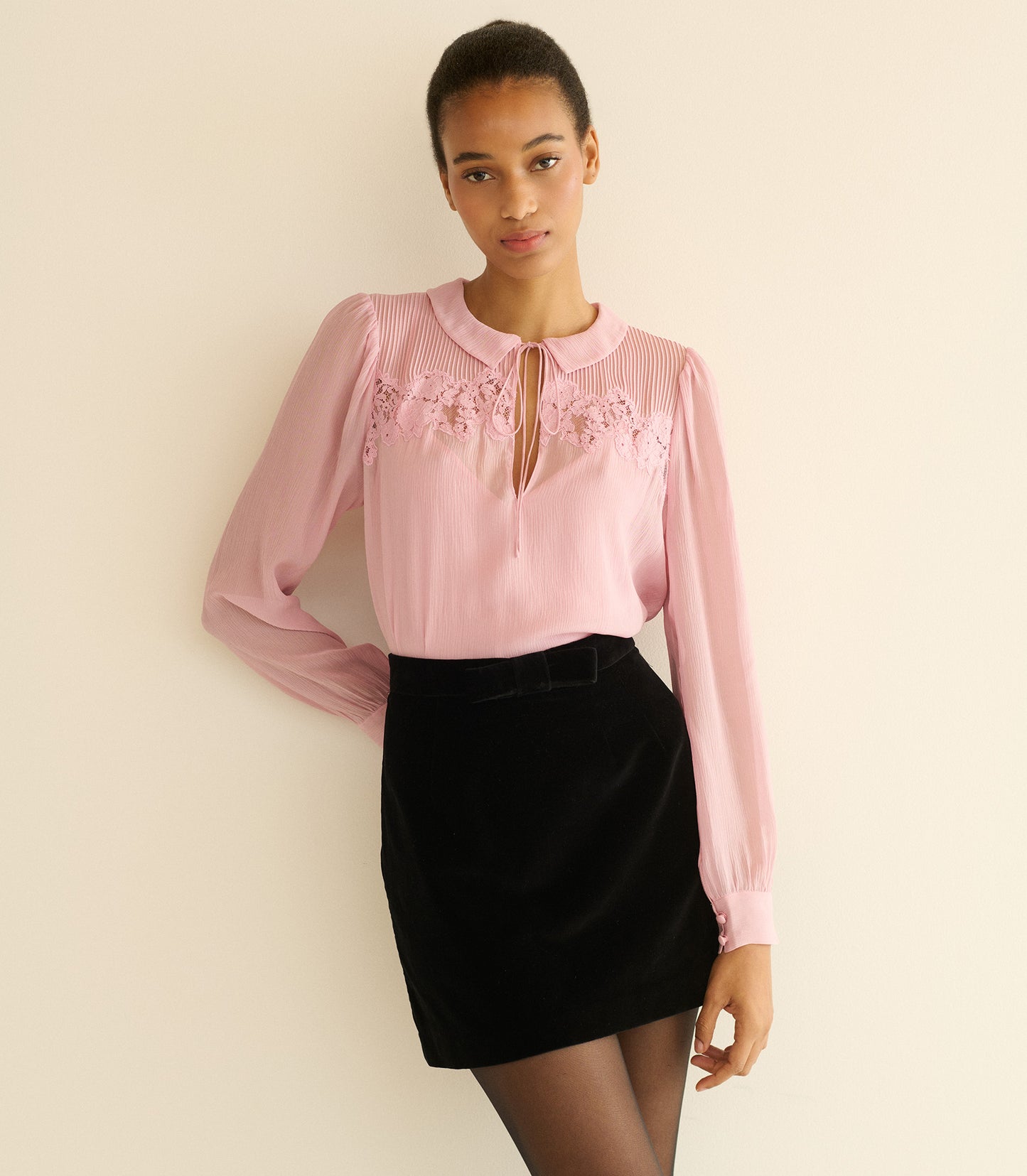 Woman wearing a pink blouse with floral details and a black skirt against a beige background
