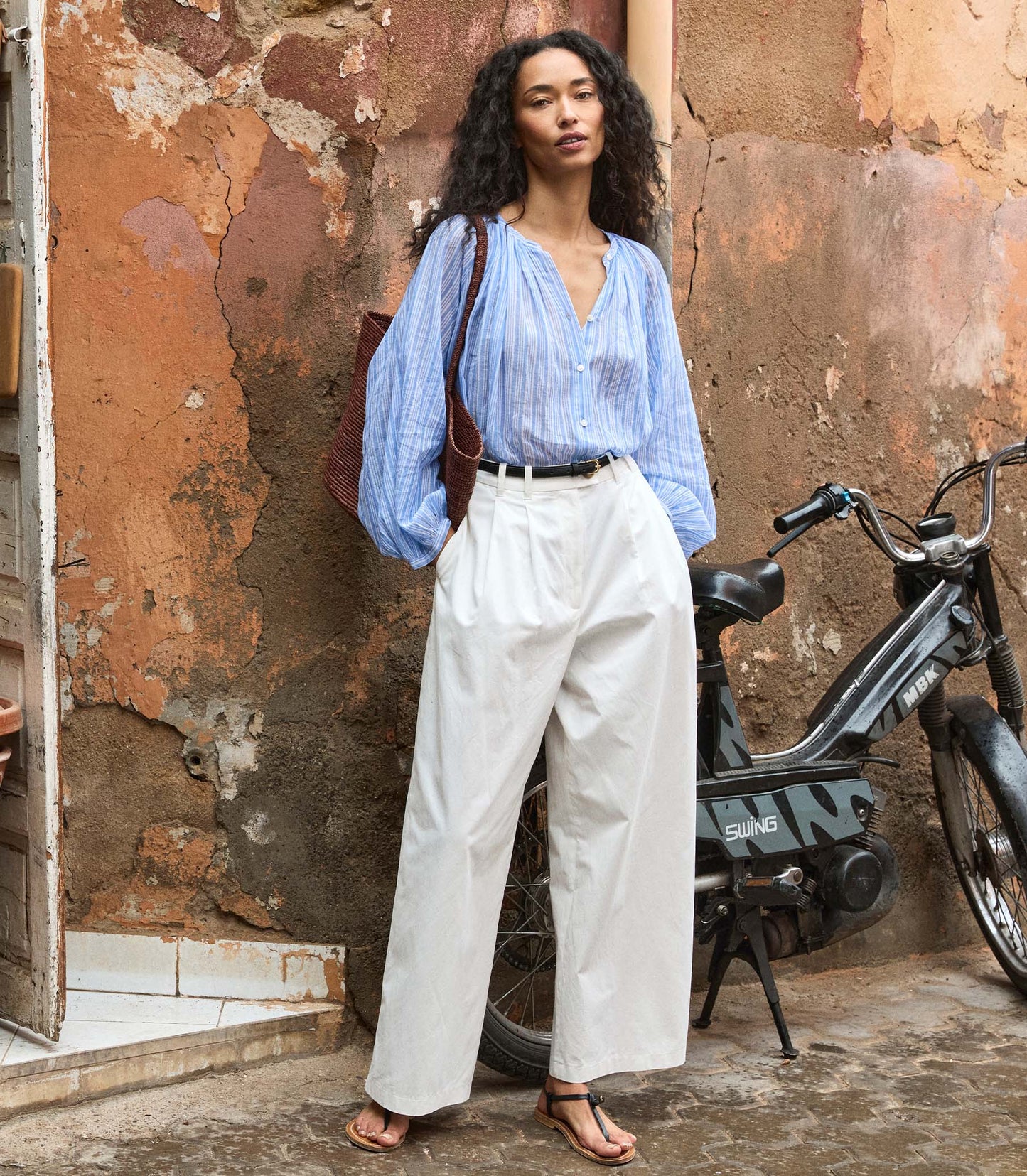 Woman in a striped blue shirt and white pants standing next to a motorcycle against a textured wall.