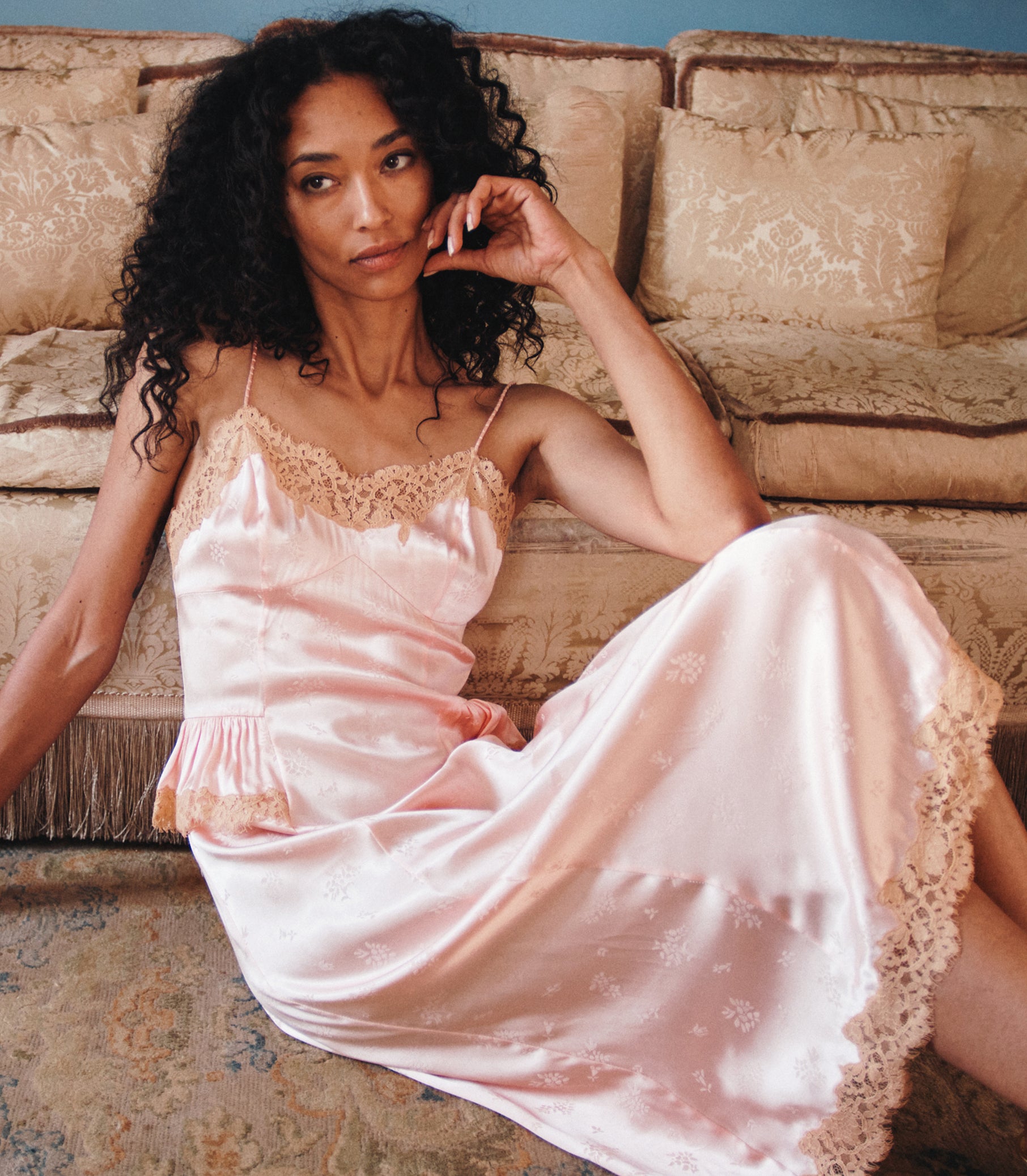 A woman with curly black hair reclines on a beige sofa wearing a light pink satin slip dress with lace trim and a subtle floral pattern. 1
