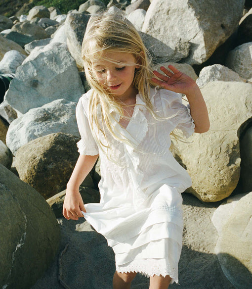 Young girl in a white blouse and white skirt sitting among large rocks.