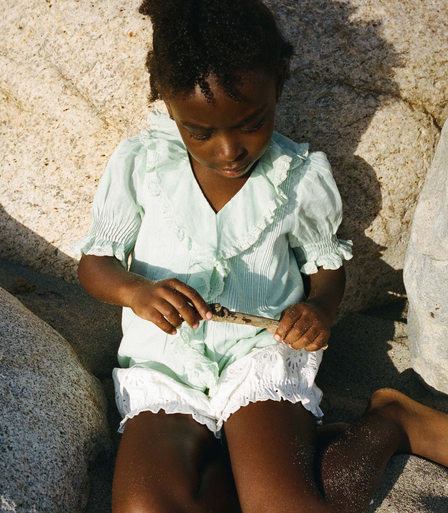 Child in a light green blouse and white bloomers sitting on rocks by a body of water.