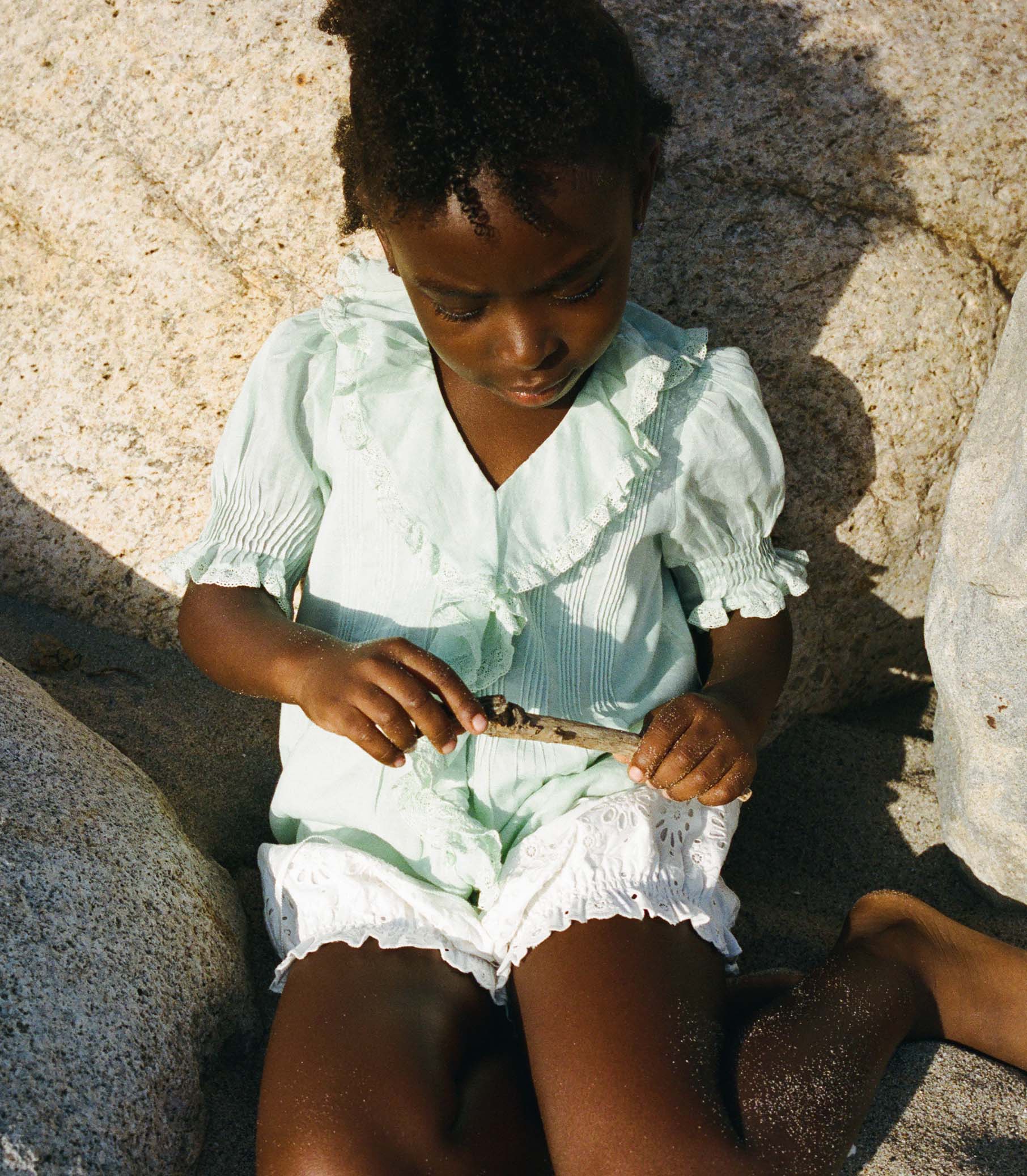 Child in a light green blouse and white bloomers sitting on rocks by a body of water. 1
