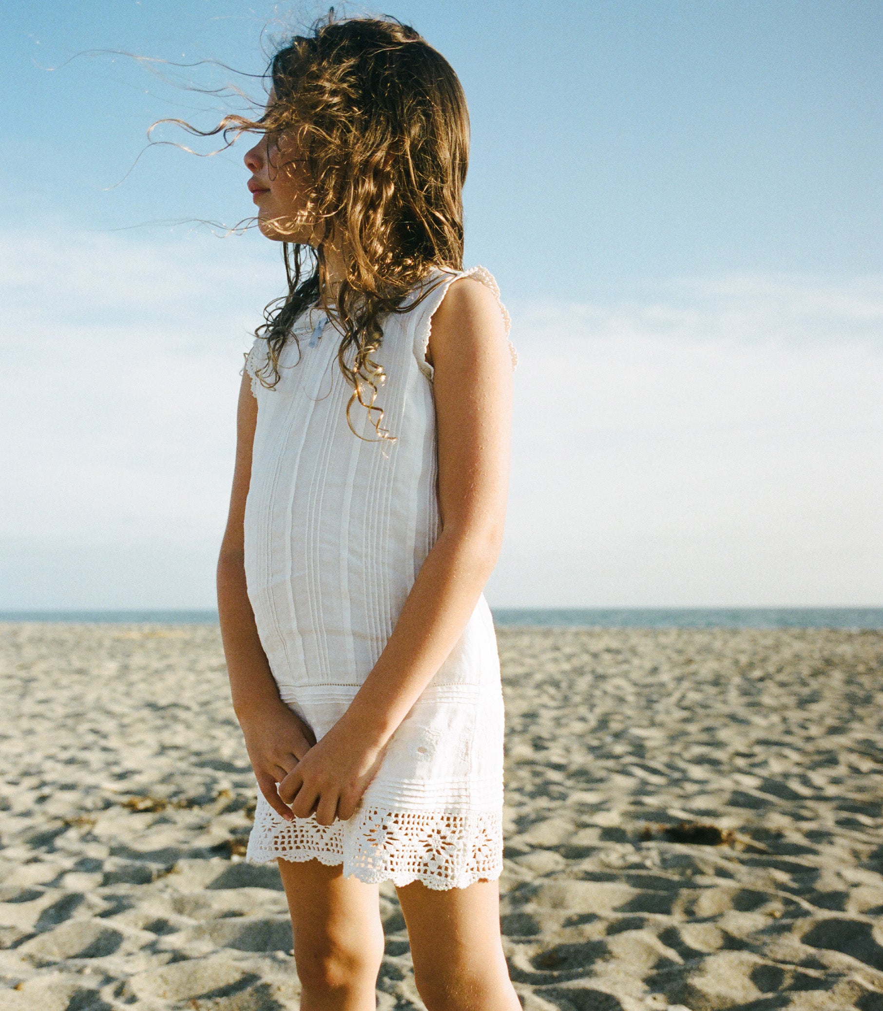 Child in a white dress standing on a sandy beach with a clear sky. 1
