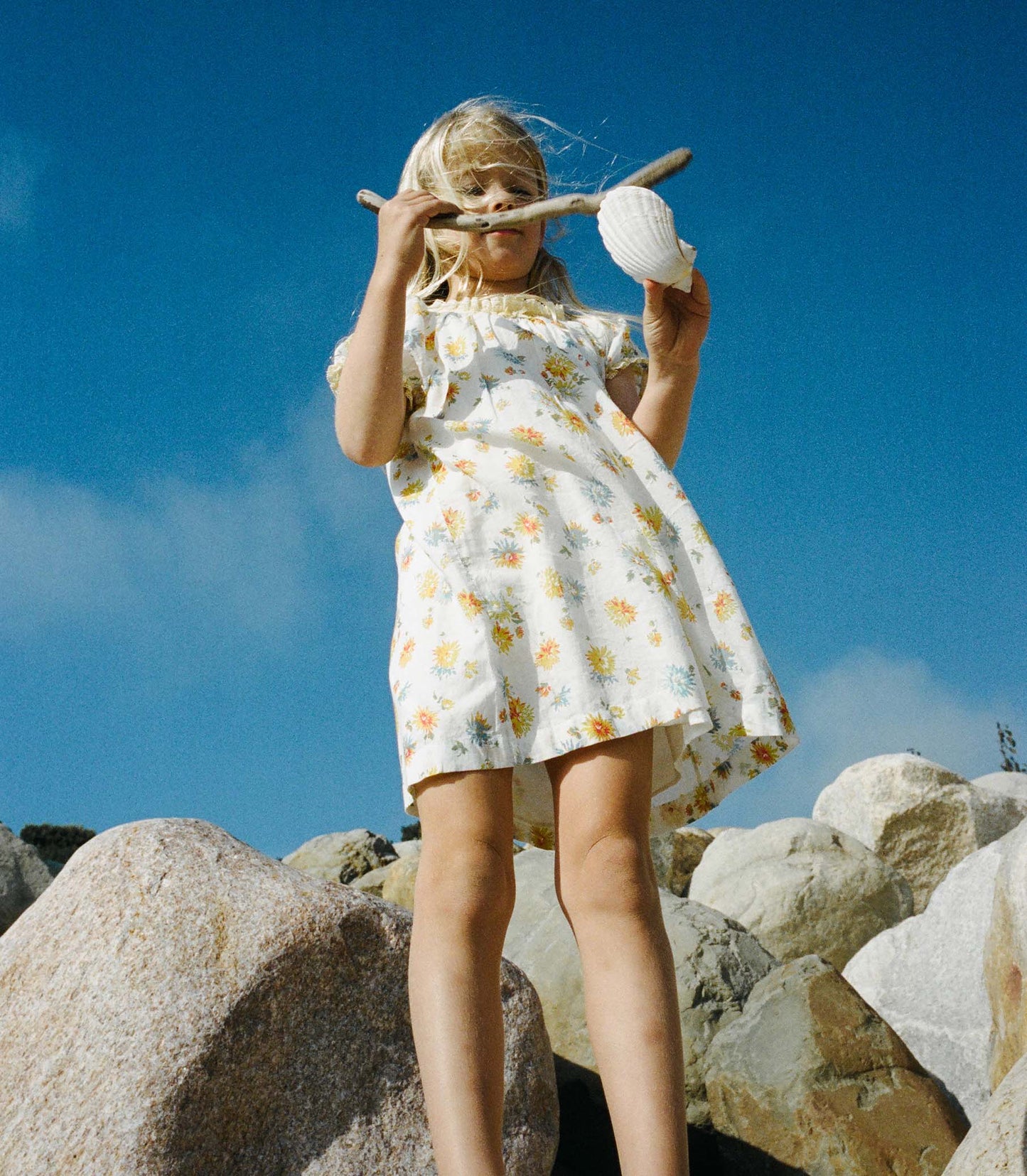 Young girl in a white floral dress holding a shell and a stick on rocky terrain with a blue sky.
