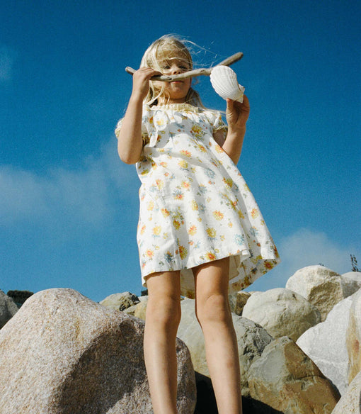 Young girl in a white floral dress holding a shell and a stick on rocky terrain with a blue sky.
