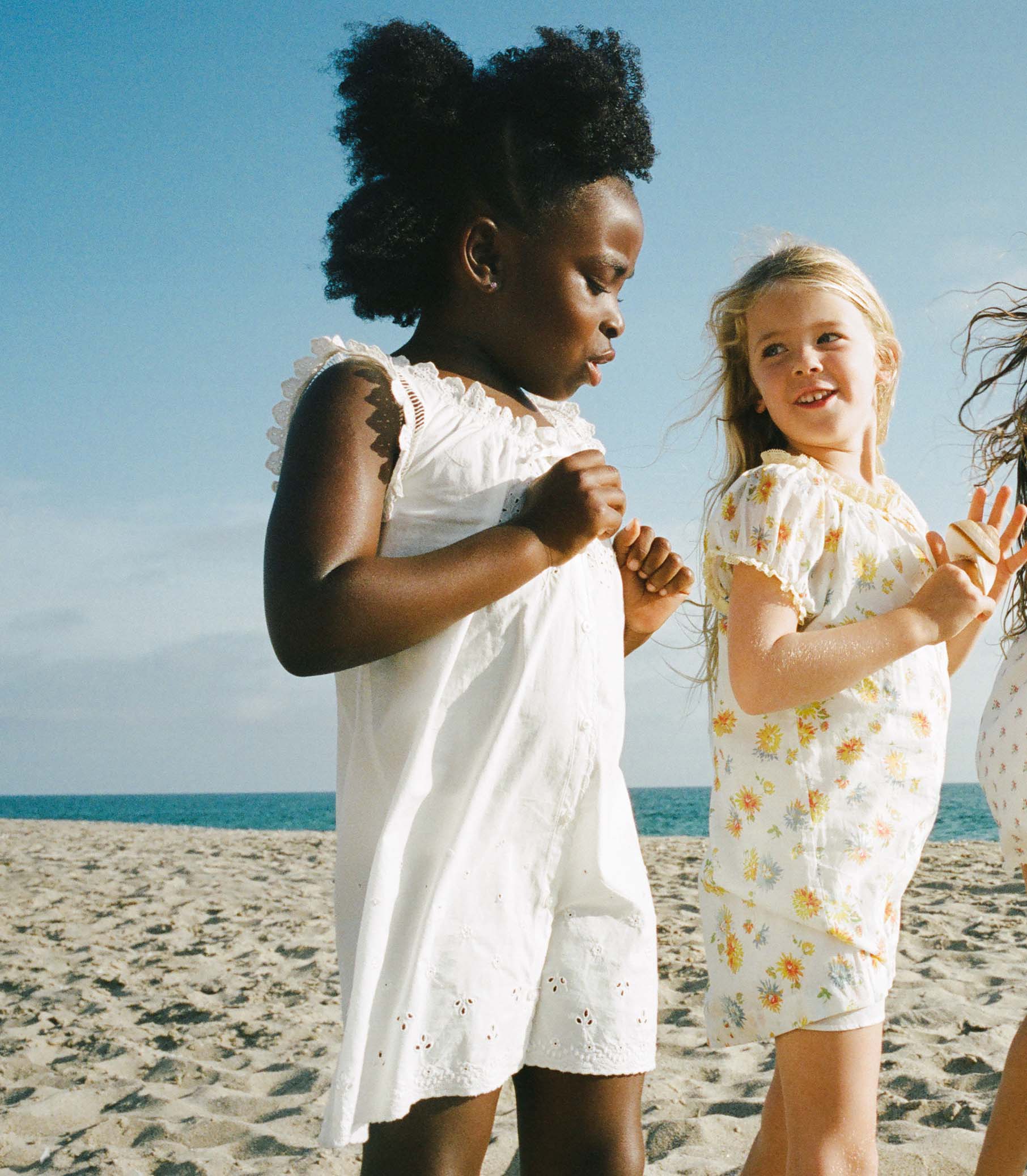 Two young girls in white dresses standing on a sandy beach with blue sky.