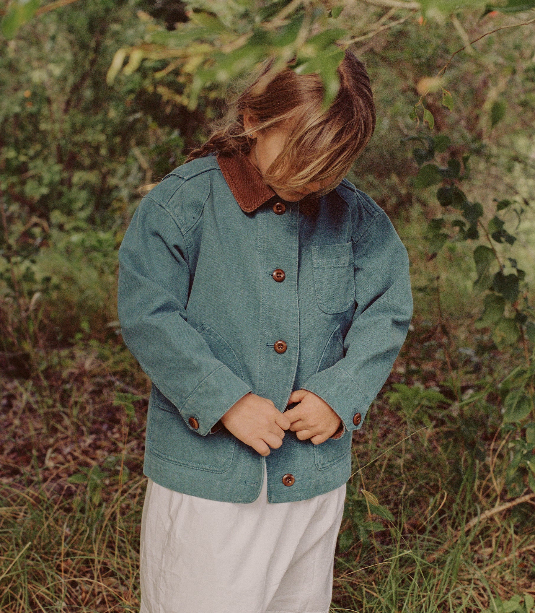 A child wearing a teal blue denim jacket with a brown corduroy collar and brown buttons, standing outdoors in a grassy, wooded area. 1

