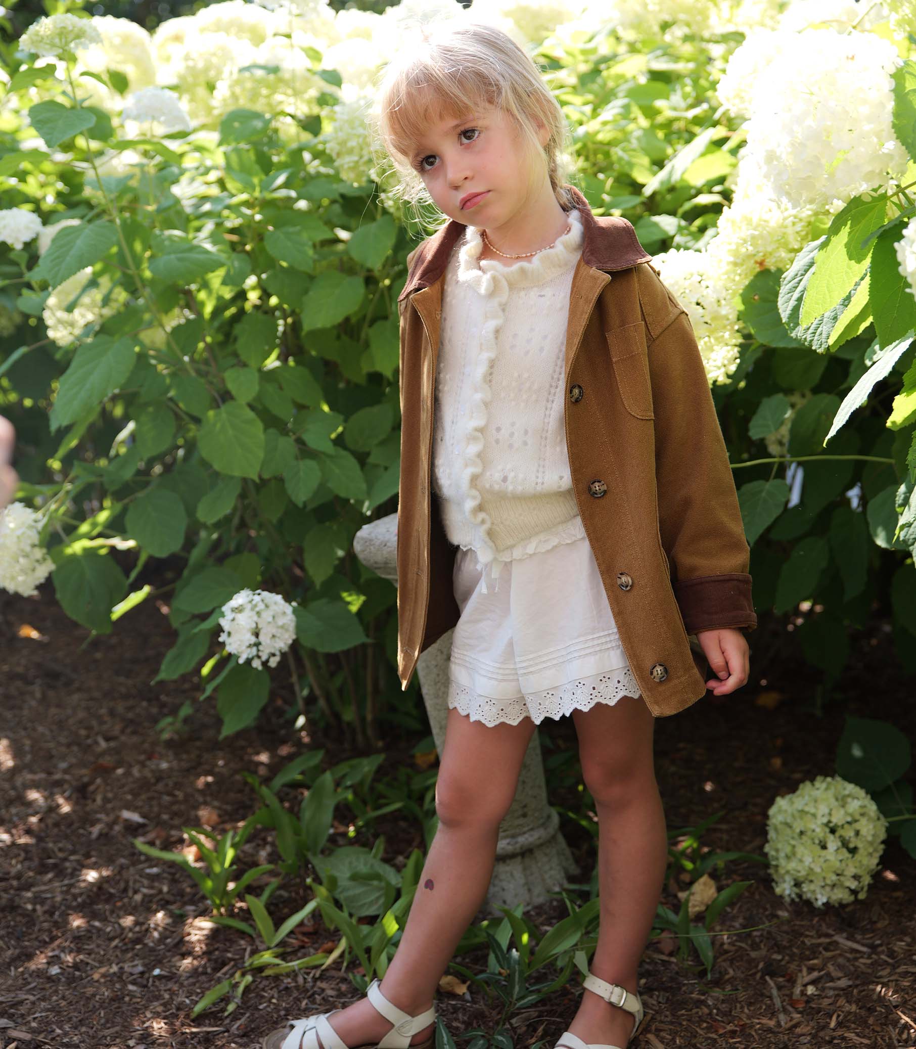 Young girl in a brown coat and white dress standing among white flowers and greenery 1