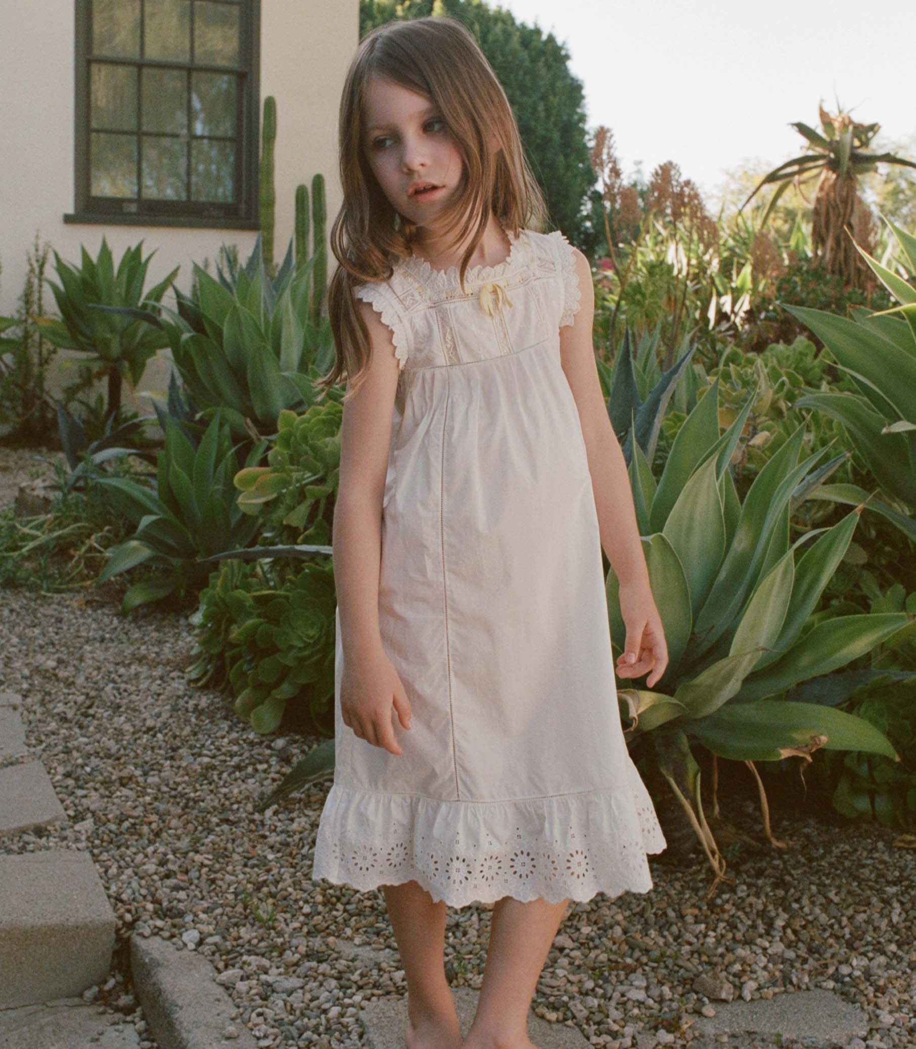 Young girl in a white dress standing in a garden with green plants and a building in the background.