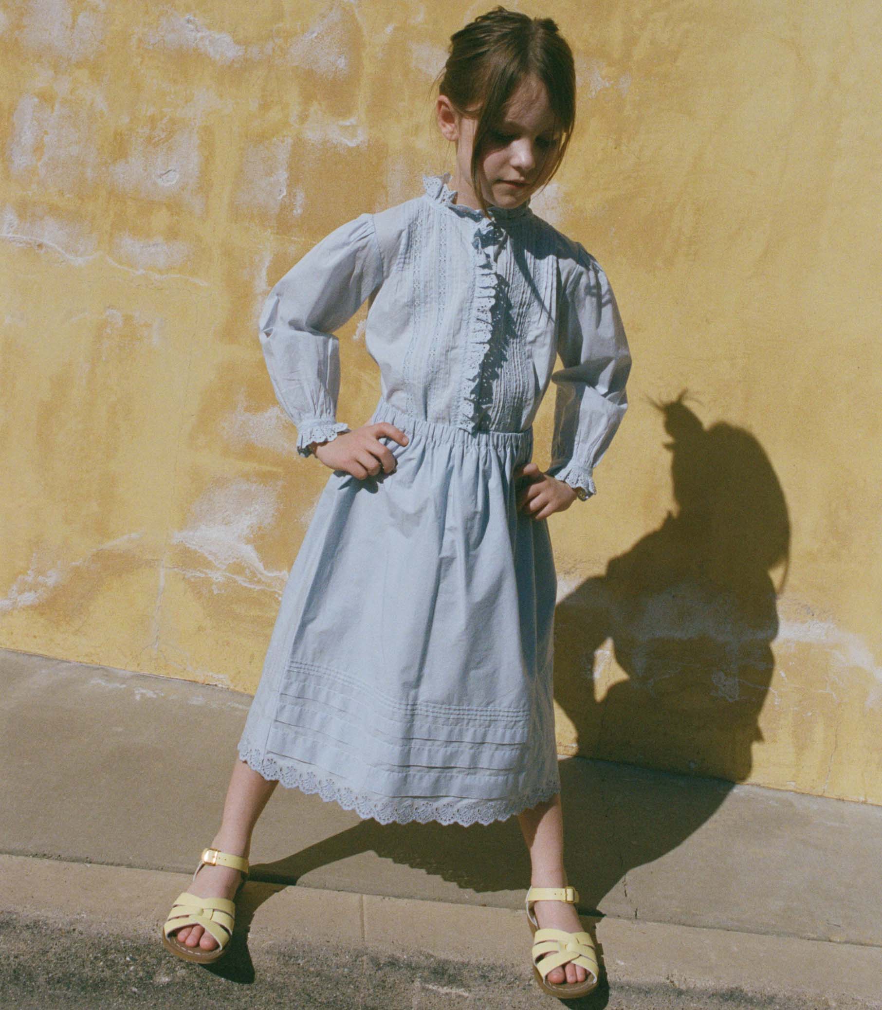 Little girl in a light blue skirt with a matching blouse standing against a yellow wall.