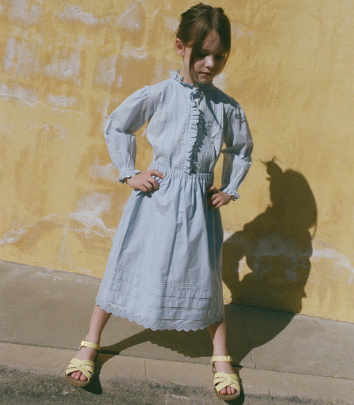 Little girl in a light blue skirt with a matching blouse standing against a yellow wall.