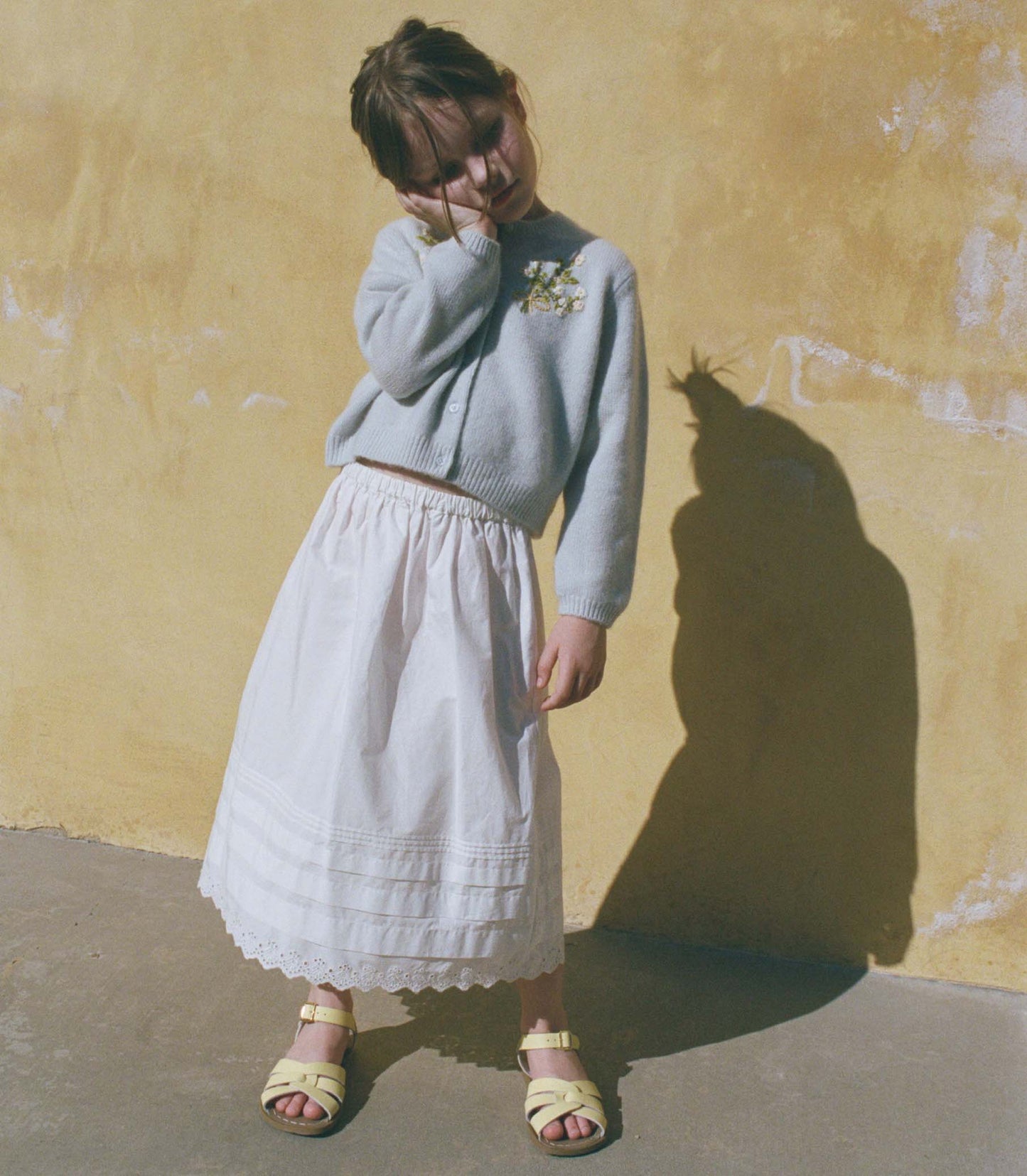Little girl wearing a light blue sweater and white skirt against a yellow wall.