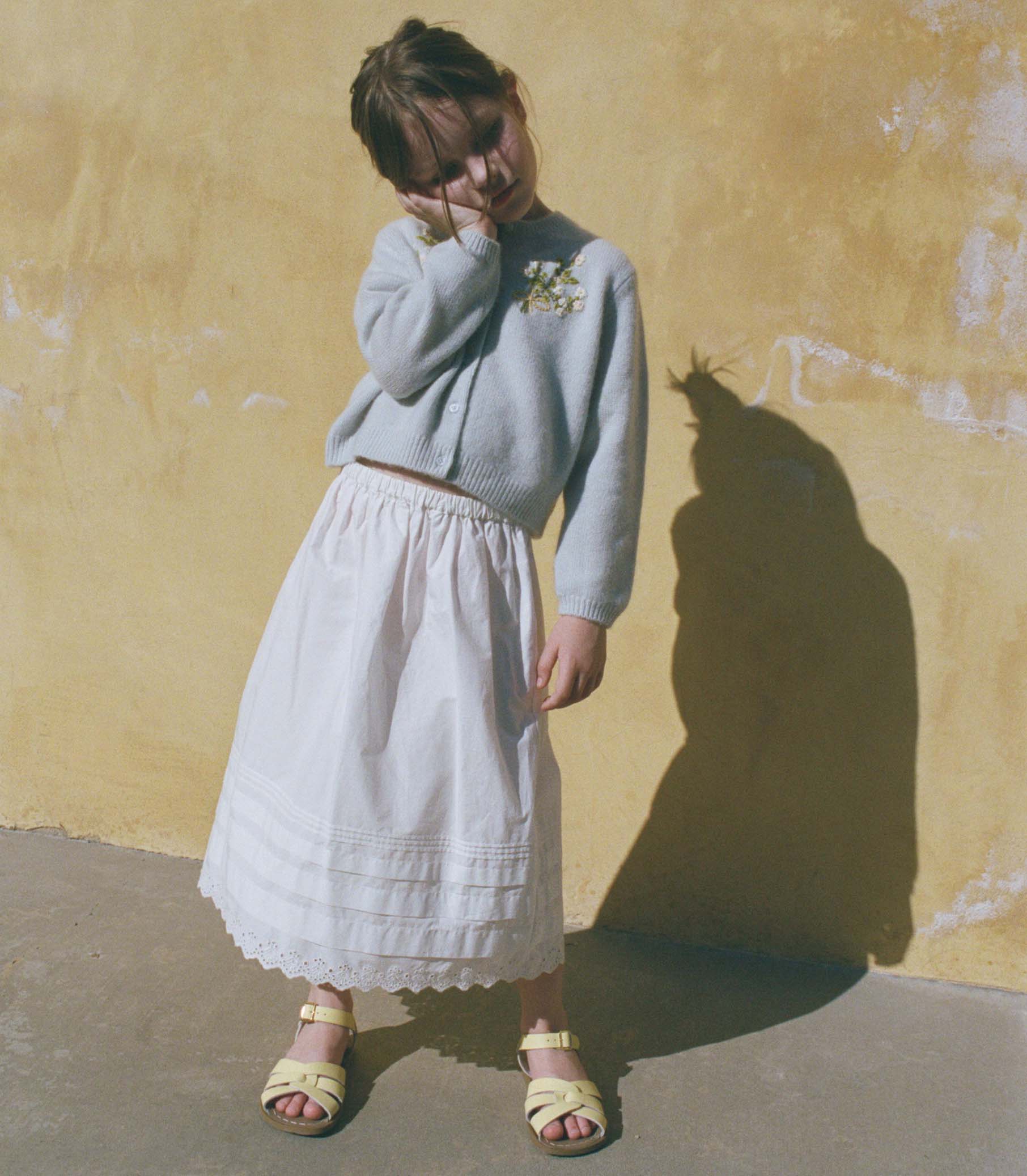 Little girl wearing a light blue sweater and white skirt against a yellow wall.