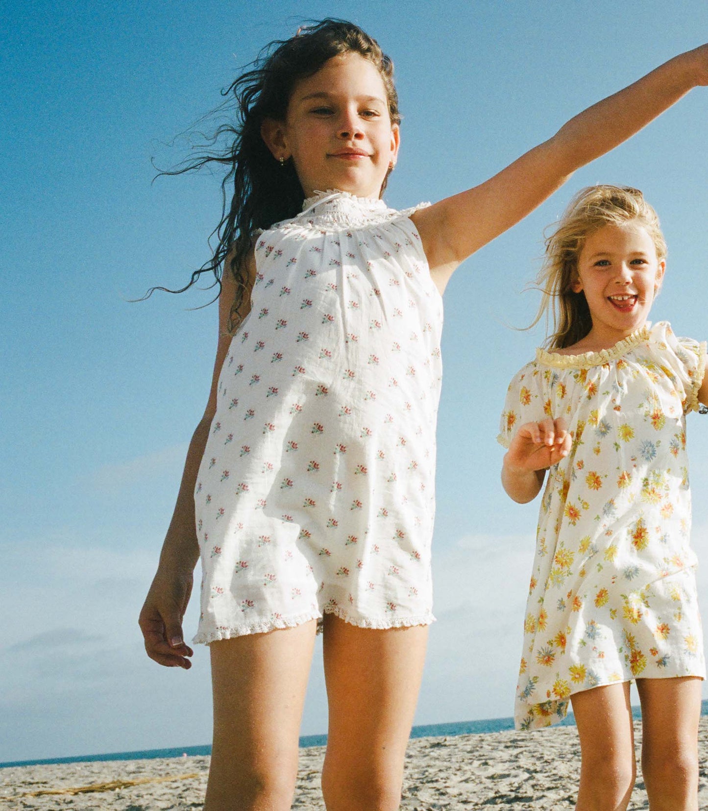 Two young girls in patterned dresses standing on a beach with a clear blue sky.