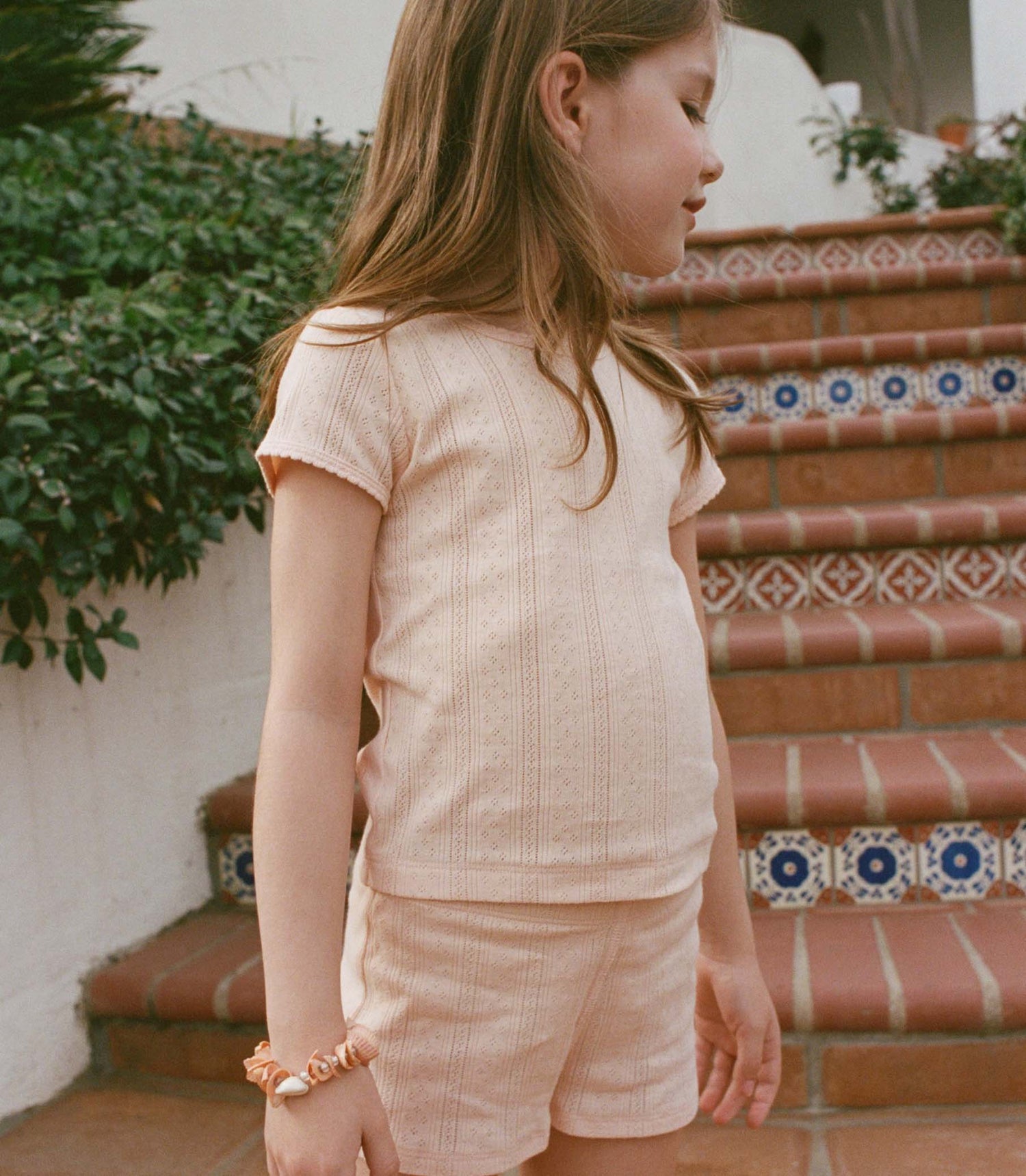 Young girl in a peach outfit standing on a tiled staircase with greenery in the background