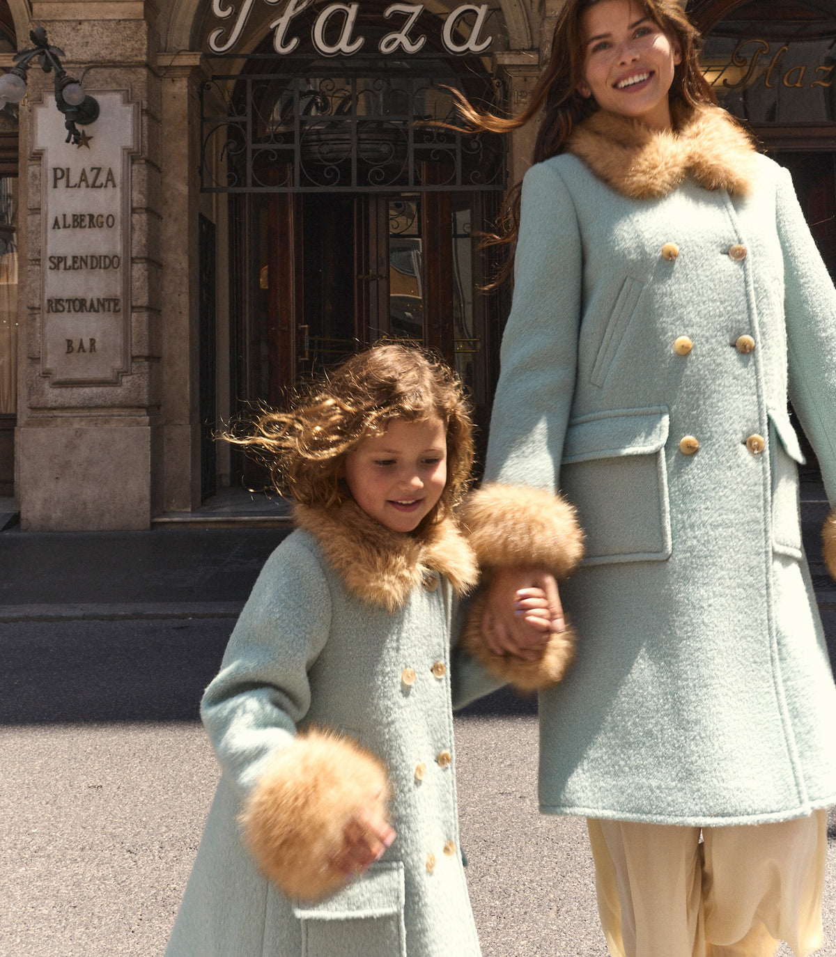 A woman and a young girl wearing matching light blue wool coats with beige fur trim and gold buttons, standing outdoors in front of a building entrance. 1
