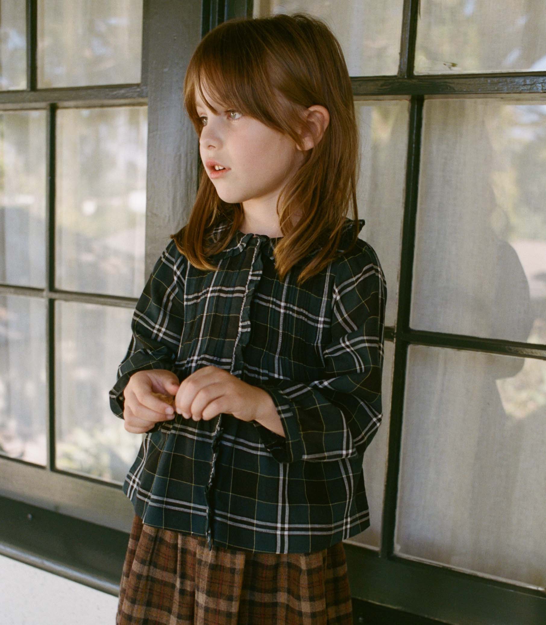 Young girl wearing a plaid shirt and skirt standing in front of a window.