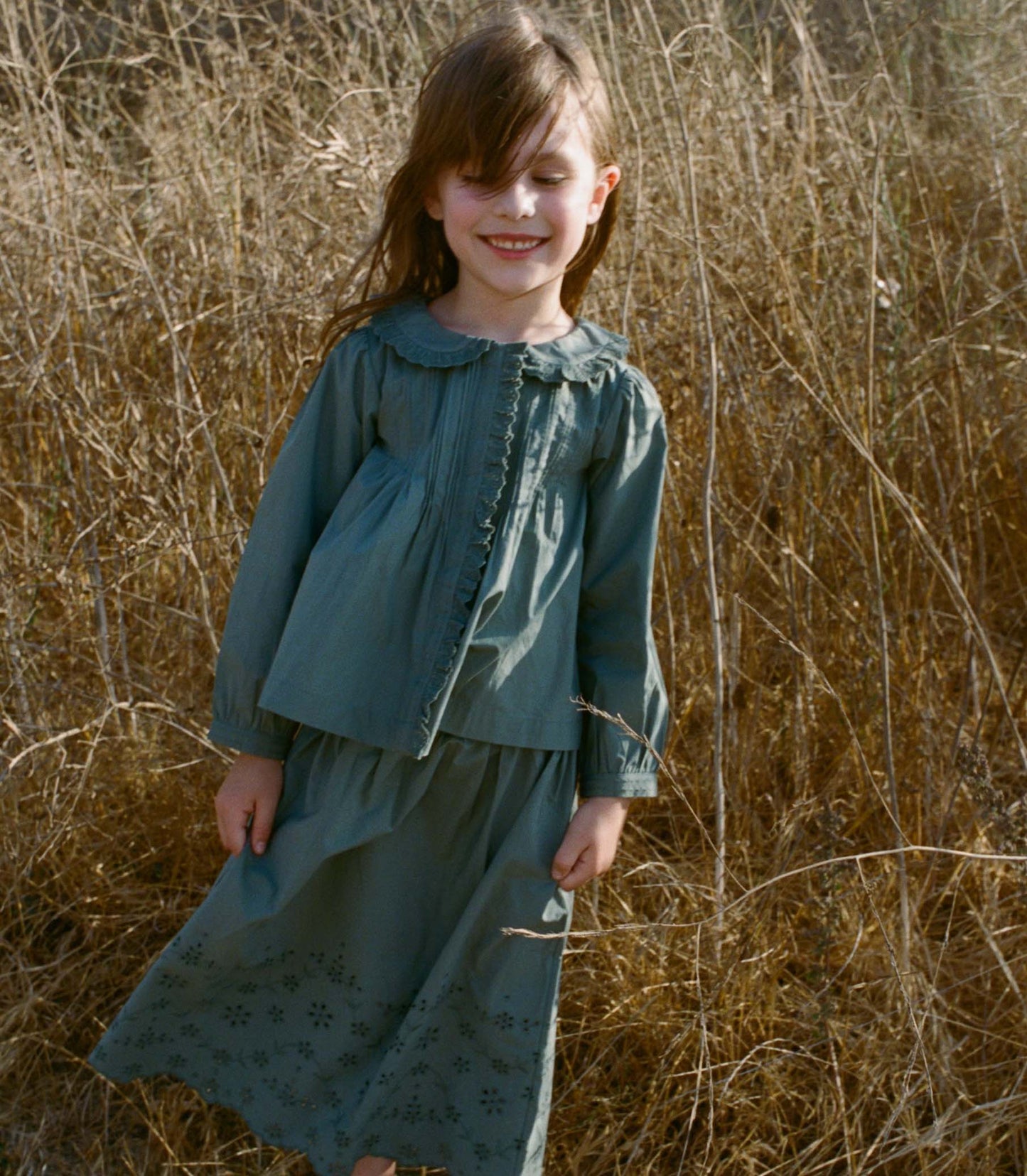 Young girl in a teal top and matching skirt, standing in a field of dry grass.