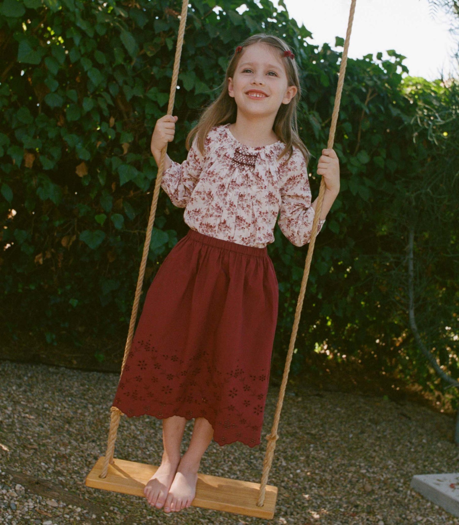 Young girl in a floral top and red skirt sitting on a wooden swing with greenery in the background.