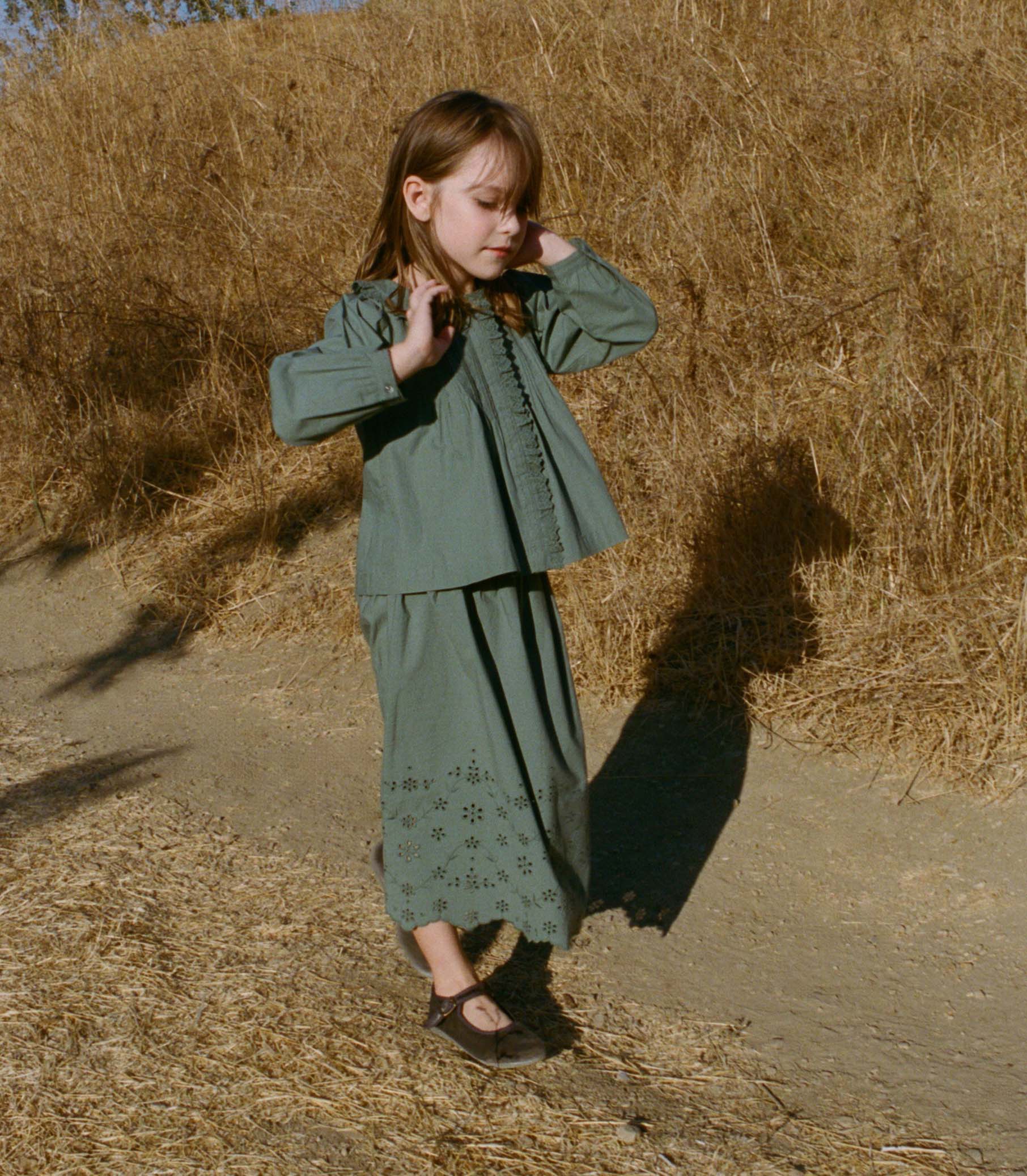 Young girl in a teal top and matching skirt, standing in a field of dry grass.