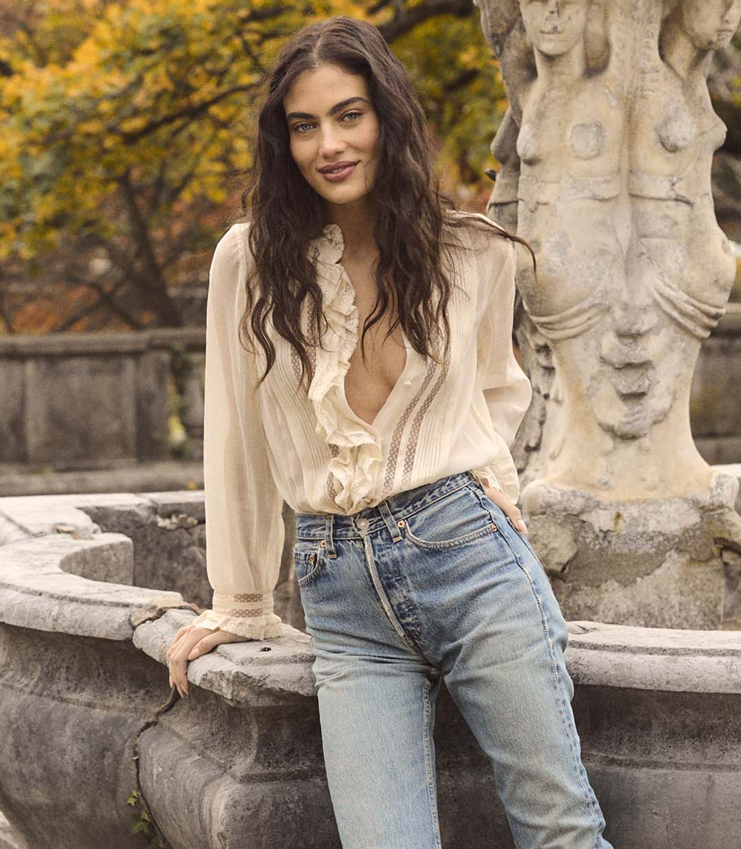 Woman in a white blouse and blue jeans leaning against a stone fountain with autumn trees in the background.