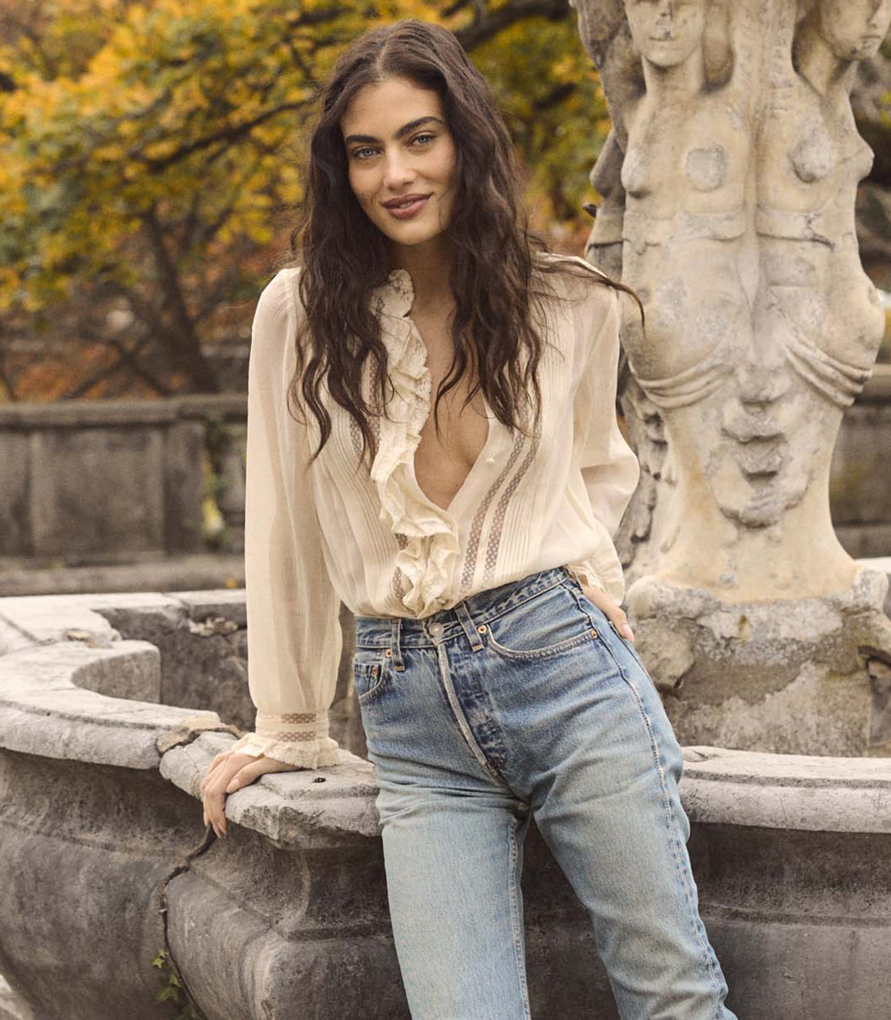 Woman in a white blouse and blue jeans leaning against a stone fountain with autumn trees in the background.