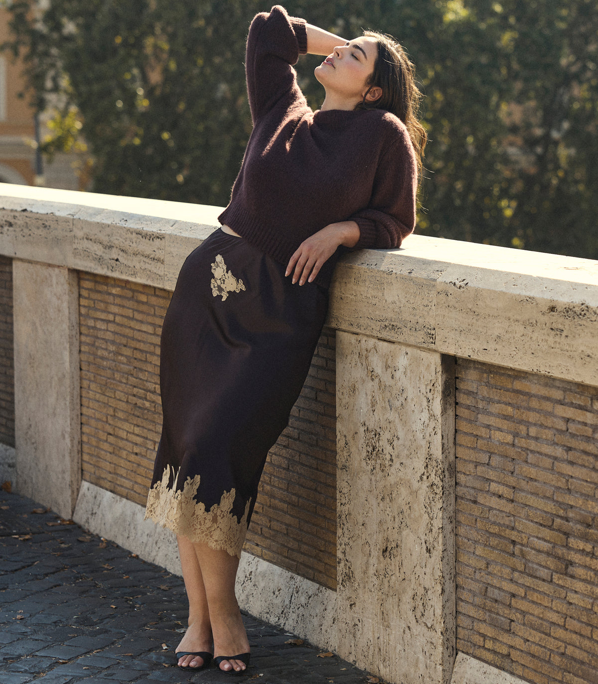 A woman wearing a dark brown knit sweater and matching midi skirt with gold lace trim leans against a stone bridge railing outdoors. 1
