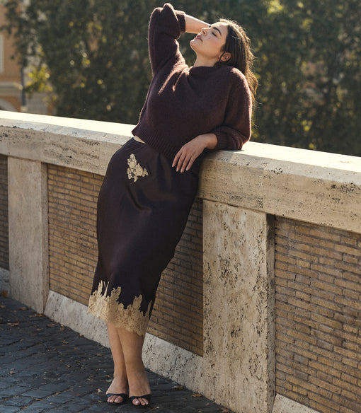 A woman wearing a dark brown knit sweater and matching midi skirt with gold lace trim leans against a stone bridge railing outdoors.