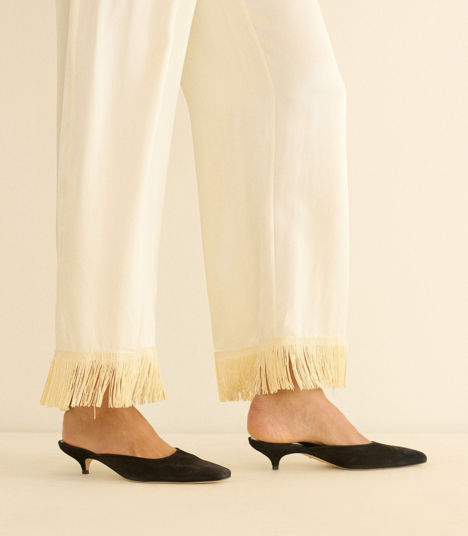 White pants with fringe at the bottom worn with black kitten heel shoes on a neutral background
