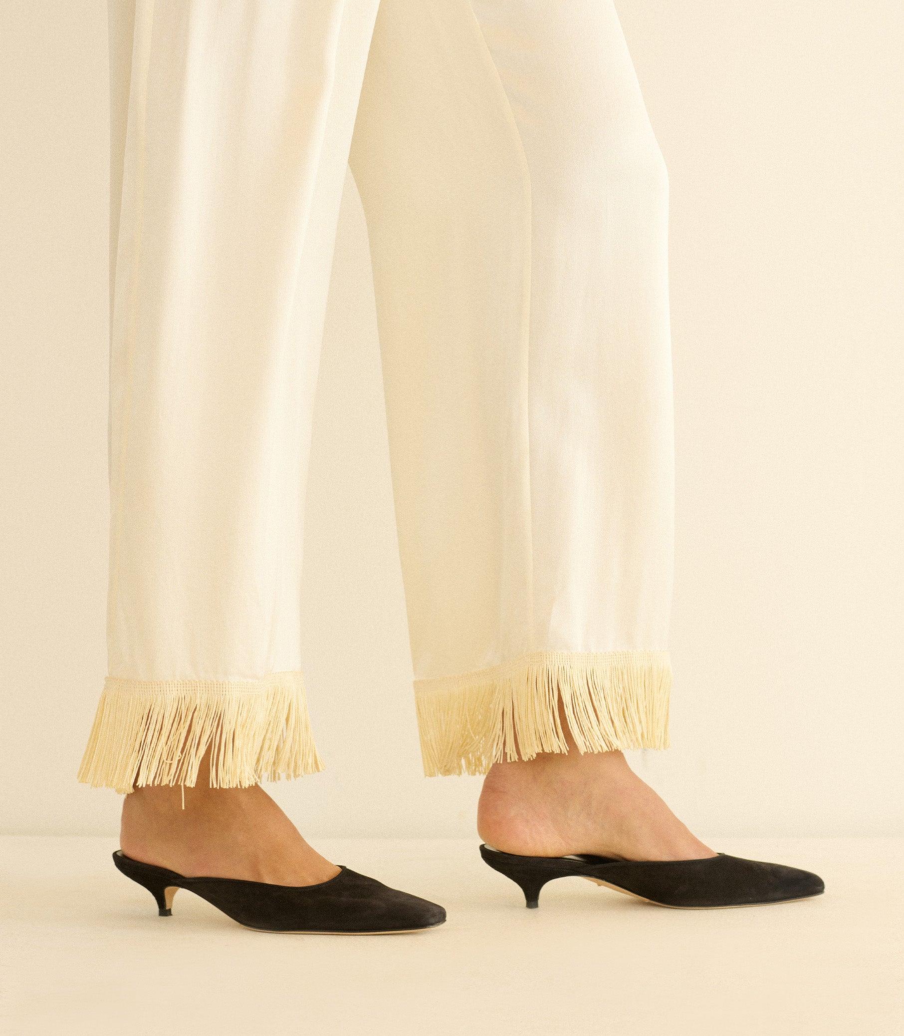White pants with fringe at the bottom worn with black kitten heel shoes on a neutral background