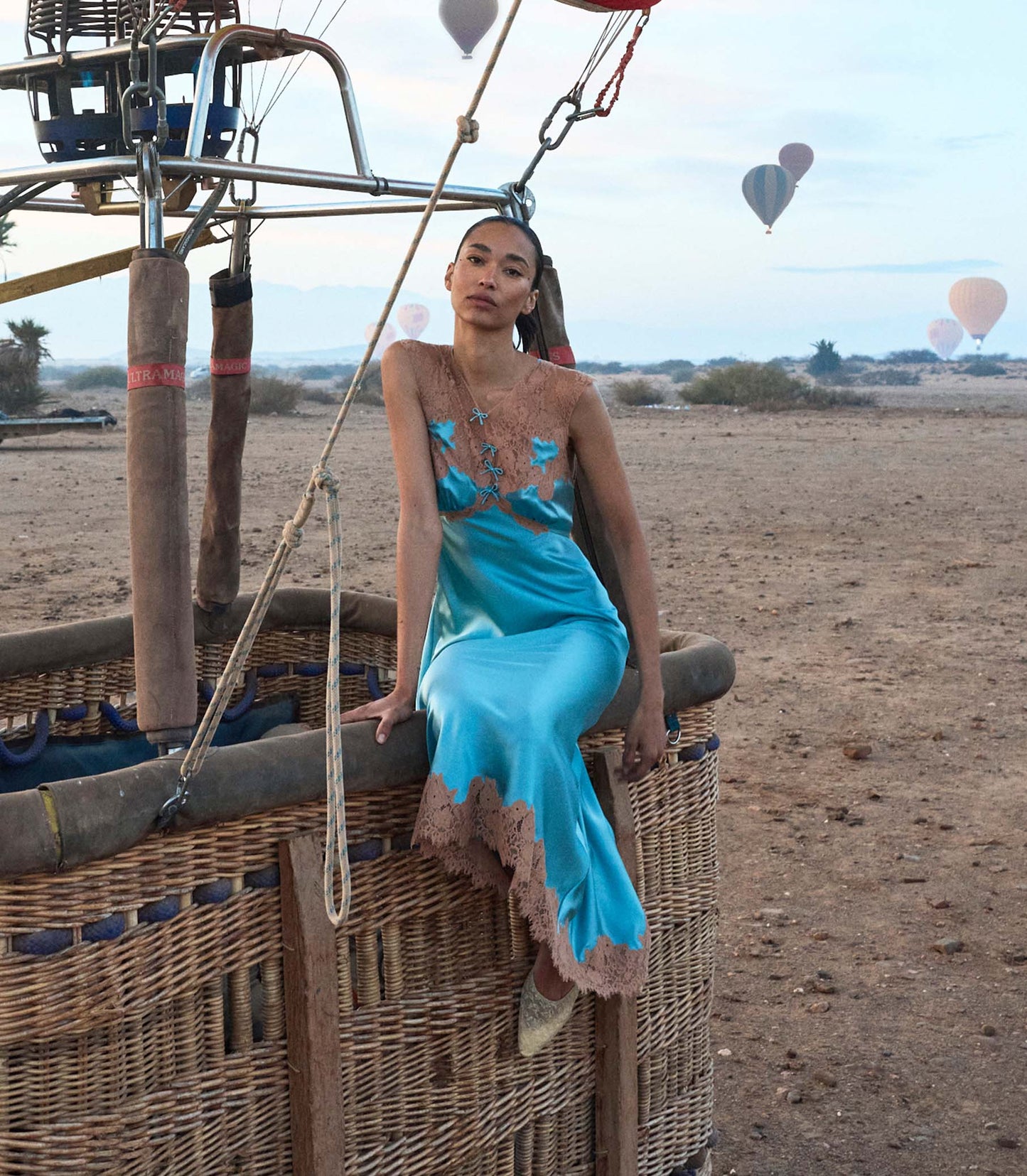 Woman in a blue silk and lace dress sitting in a hot air balloon basket with other balloons in the background.
