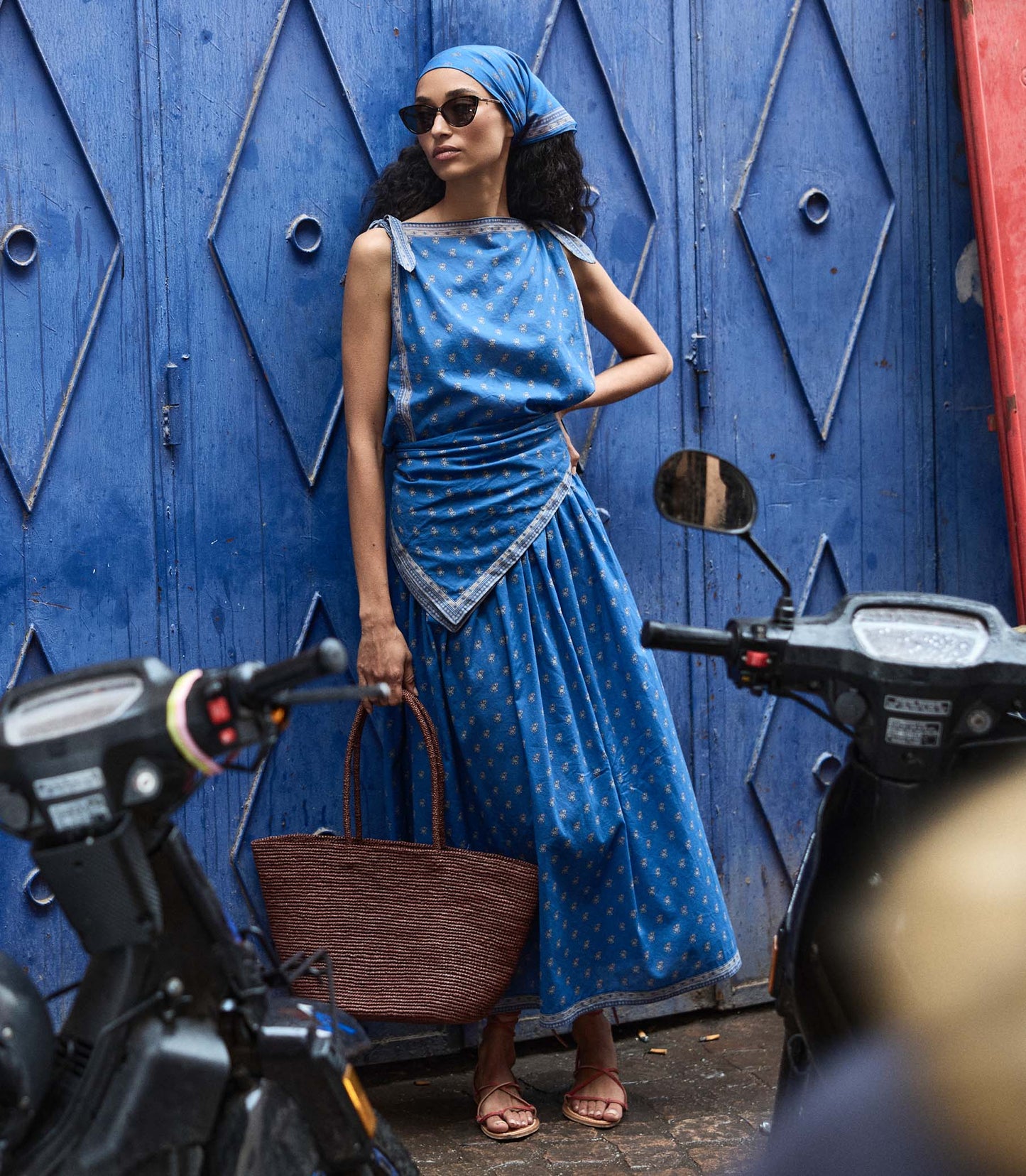 Woman in a blue paisley skirt and matching top standing next to a motorcycle with a blue wall in the background