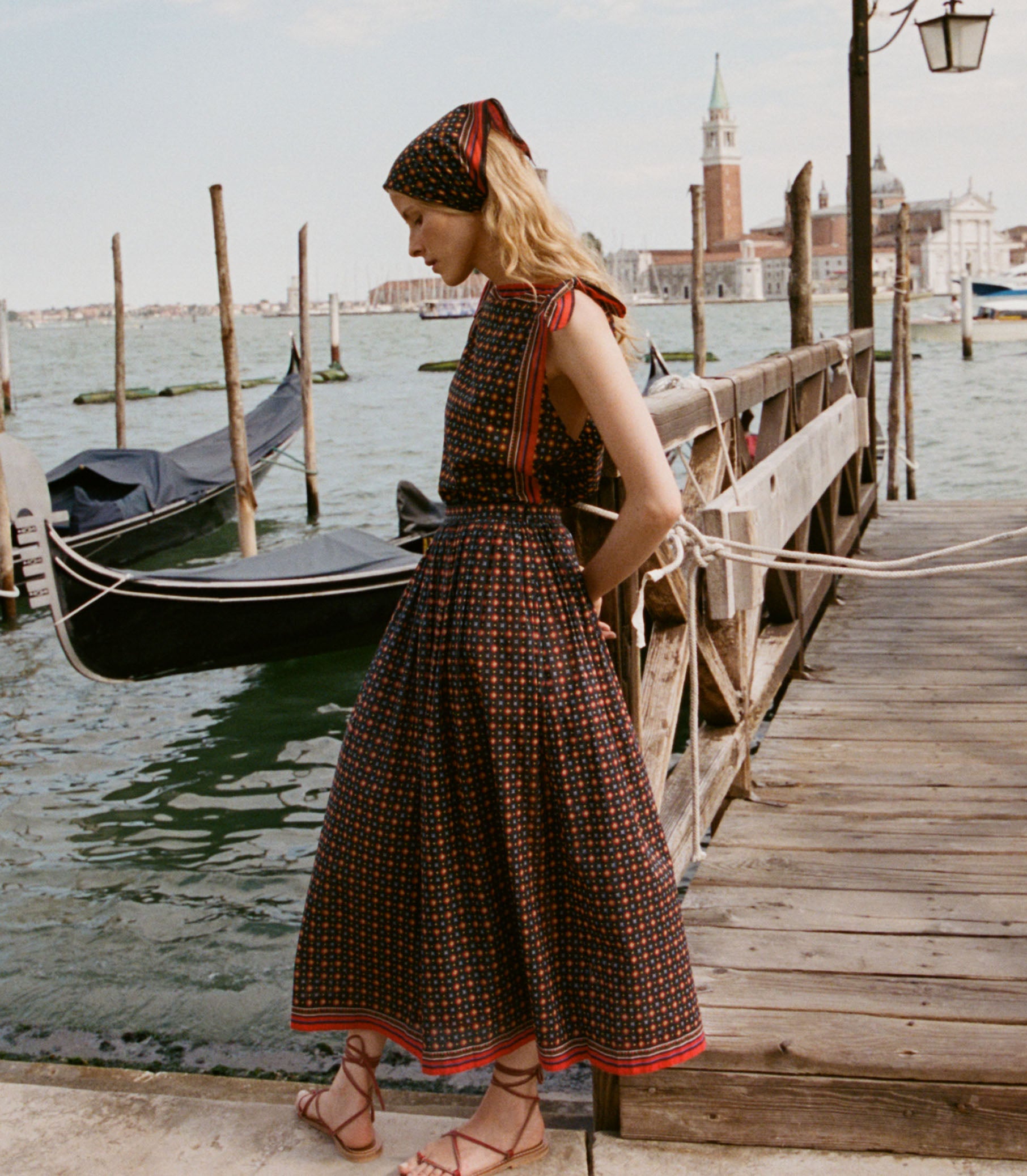 Woman in a patterned dress standing on a dock with gondolas and a cityscape in the background