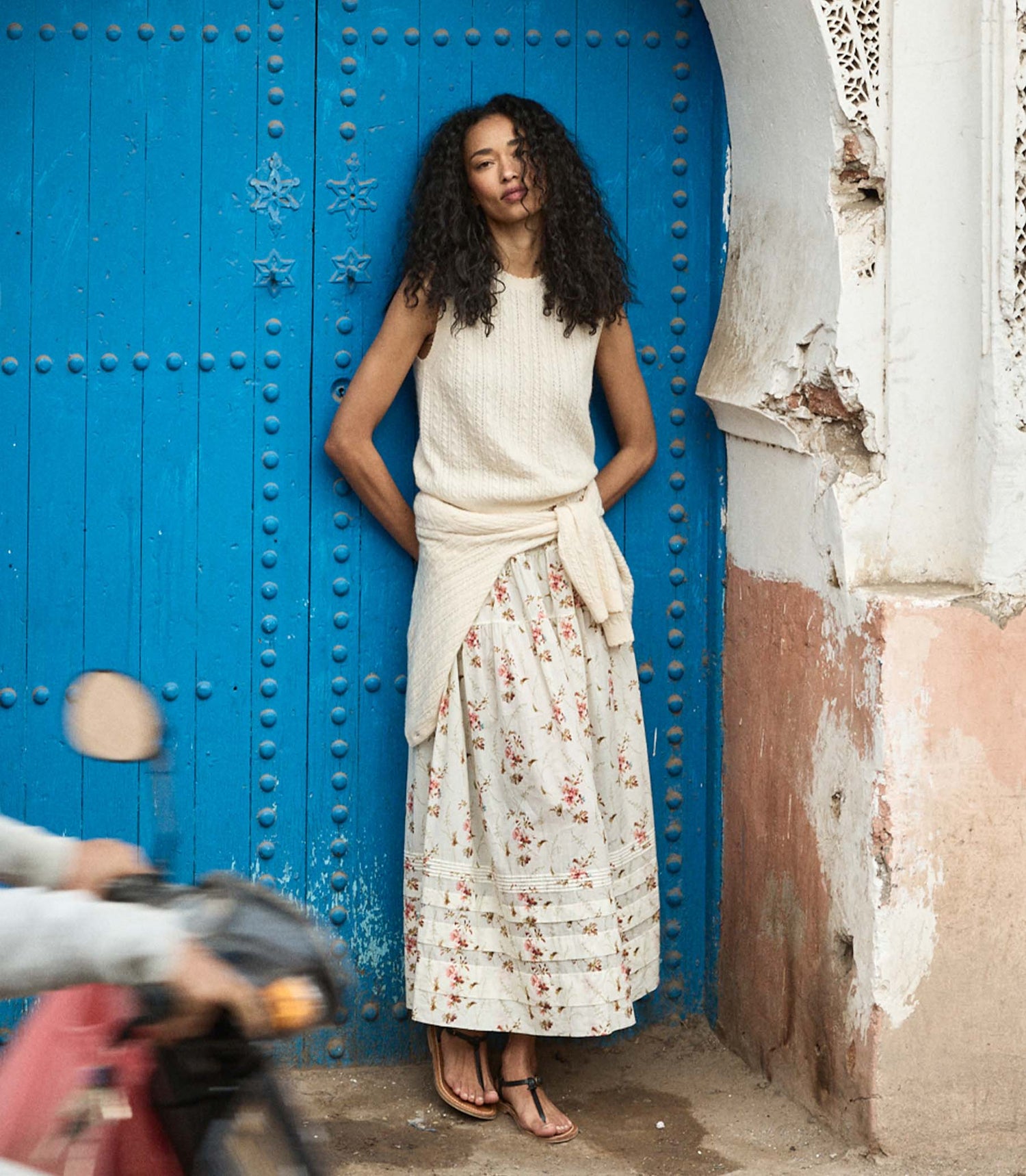 Woman in a white top with floral skirt standing in front of a blue door.