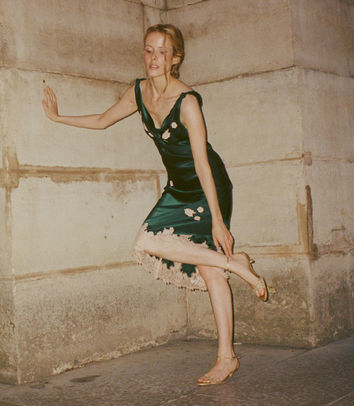 A woman in a dark green satin dress with white floral embroidery and a ruffled hem poses dynamically against a textured stone wall. 1
