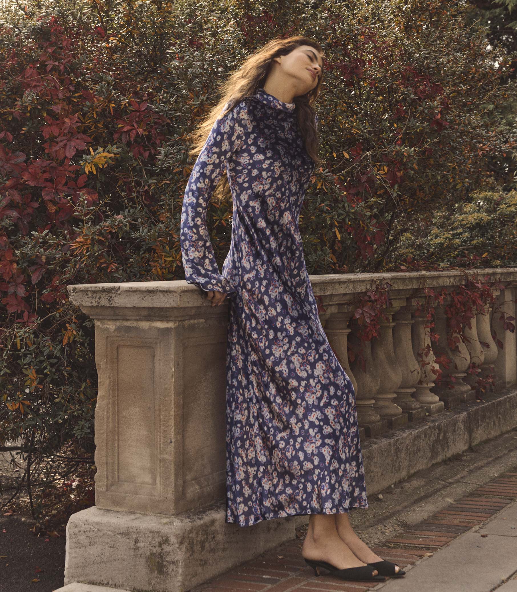 Woman in a purple floral dress standing against a stone wall with foliage in the background 1