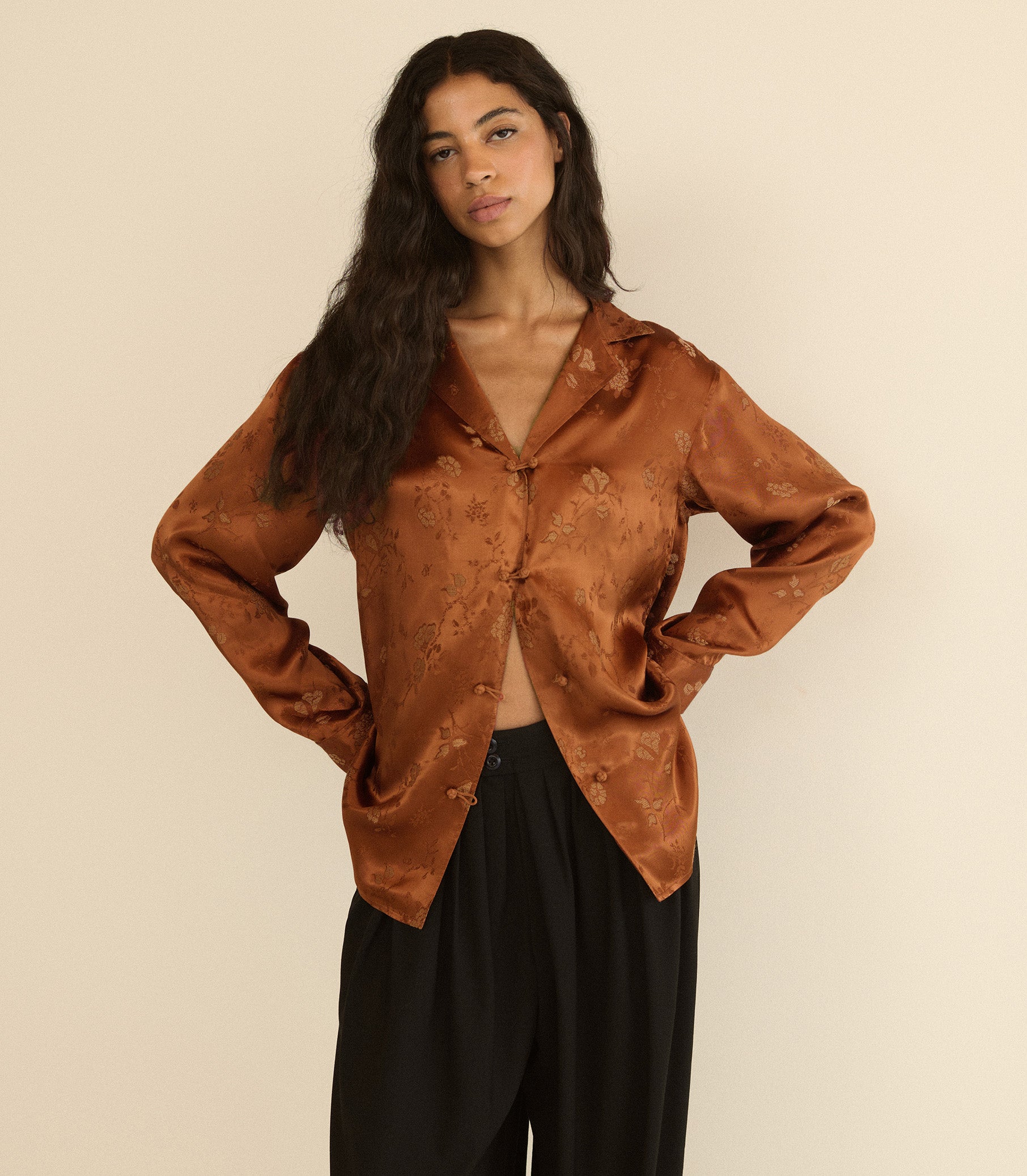 A woman modeling a long-sleeve satin-like button-front blouse in a warm rust brown with a subtle tonal floral jacquard pattern, paired with black pants, against a plain beige studio background. 1
