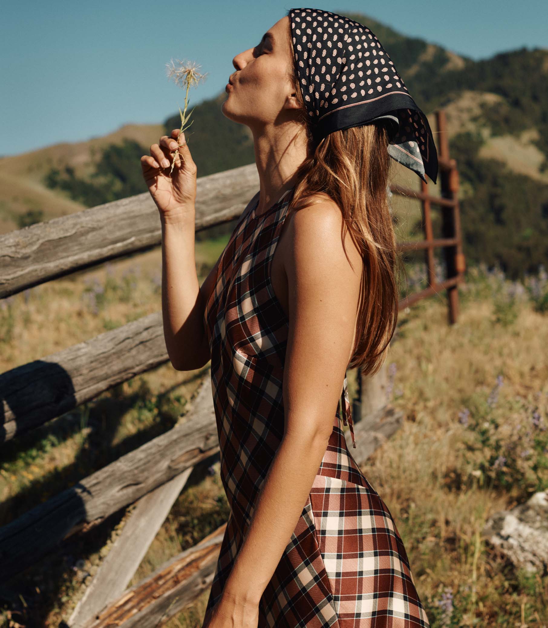 Woman in a plaid dress and patterned headscarf holding a flower in a natural setting with mountains.