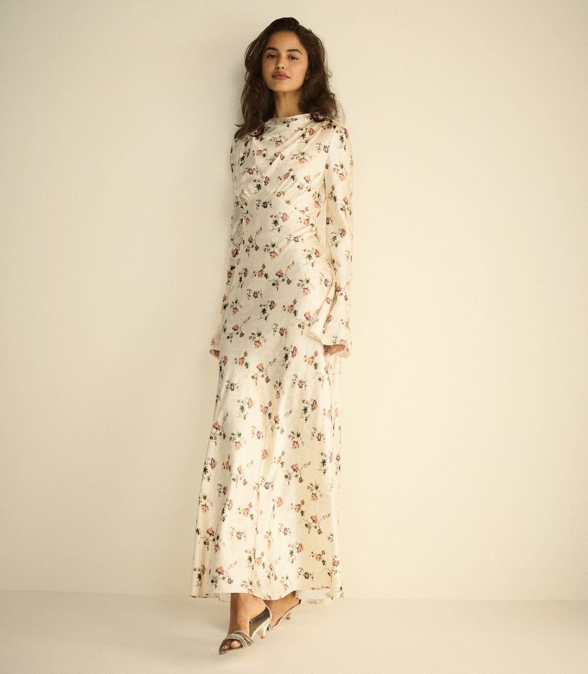 A woman modeling a long-sleeved, high-neck maxi dress with a delicate floral print on a cream background, standing against a plain beige studio backdrop.
