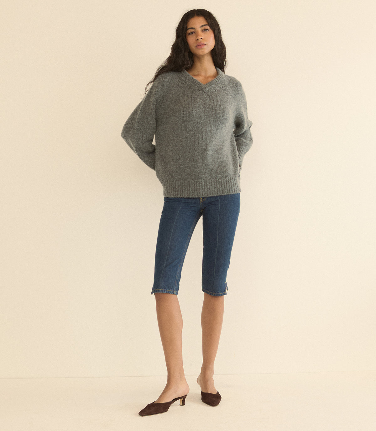 A woman modeling a gray knit V-neck sweater and blue cropped jeans against a plain beige studio background. 1
