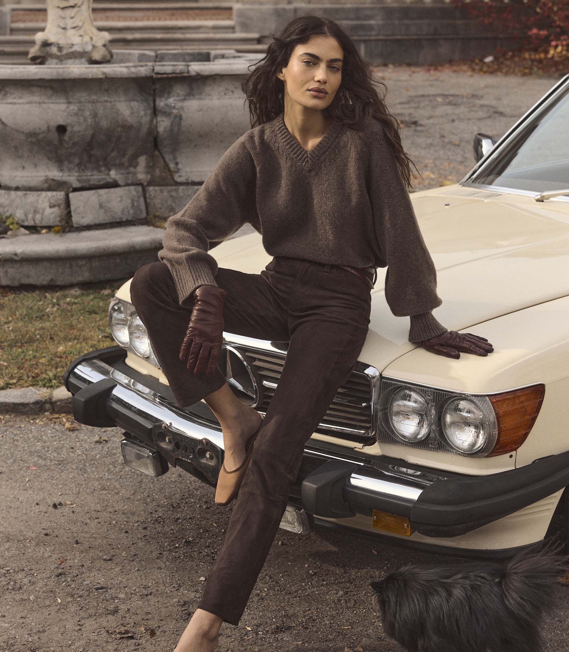 Woman wearing a brown sweater and brown pants, sitting on a vintage car in an urban setting 1