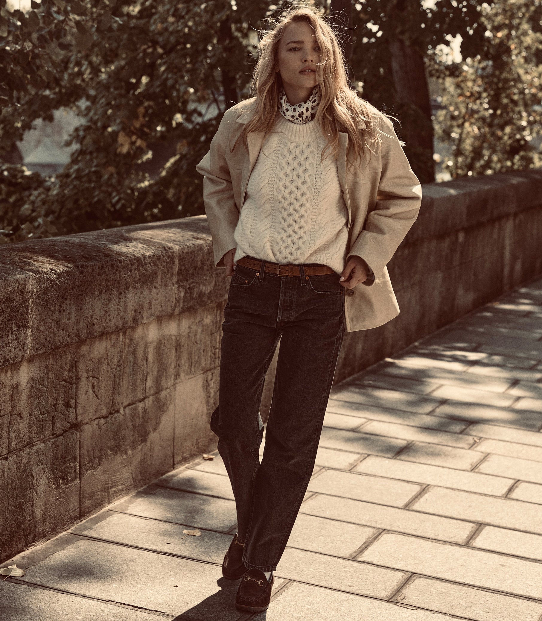 A woman stands on a stone walkway outdoors wearing a beige trench coat over a cream cable-knit sweater, dark jeans, and brown shoes. 1
