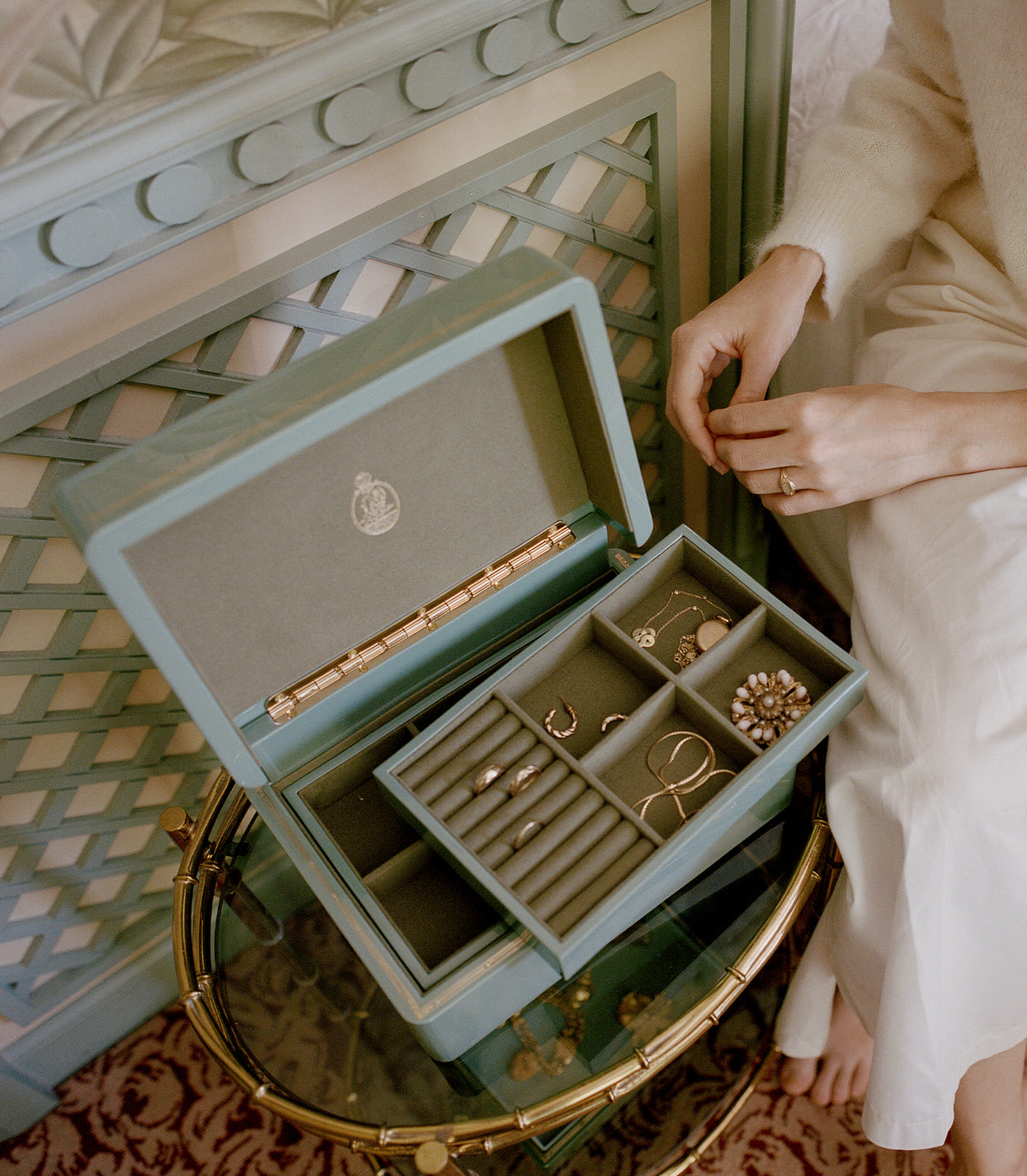 An open sage green jewelry box with multiple compartments, styled on a bamboo-style side table next to a person wearing a cream sweater and white dress. 1
