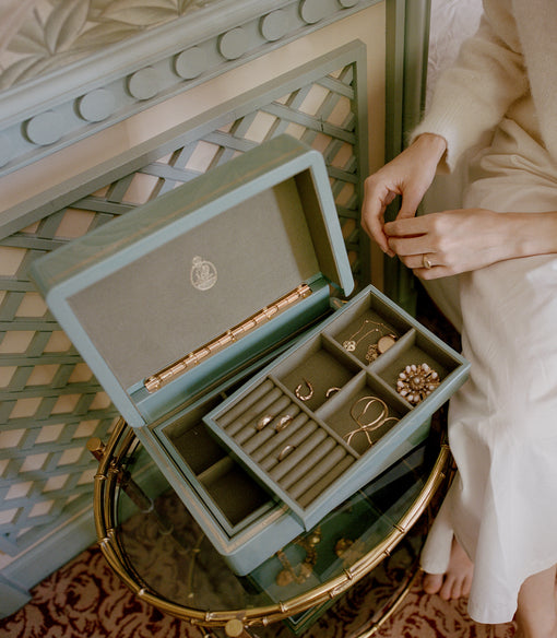 An open sage green jewelry box with multiple compartments, styled on a bamboo-style side table next to a person wearing a cream sweater and white dress.