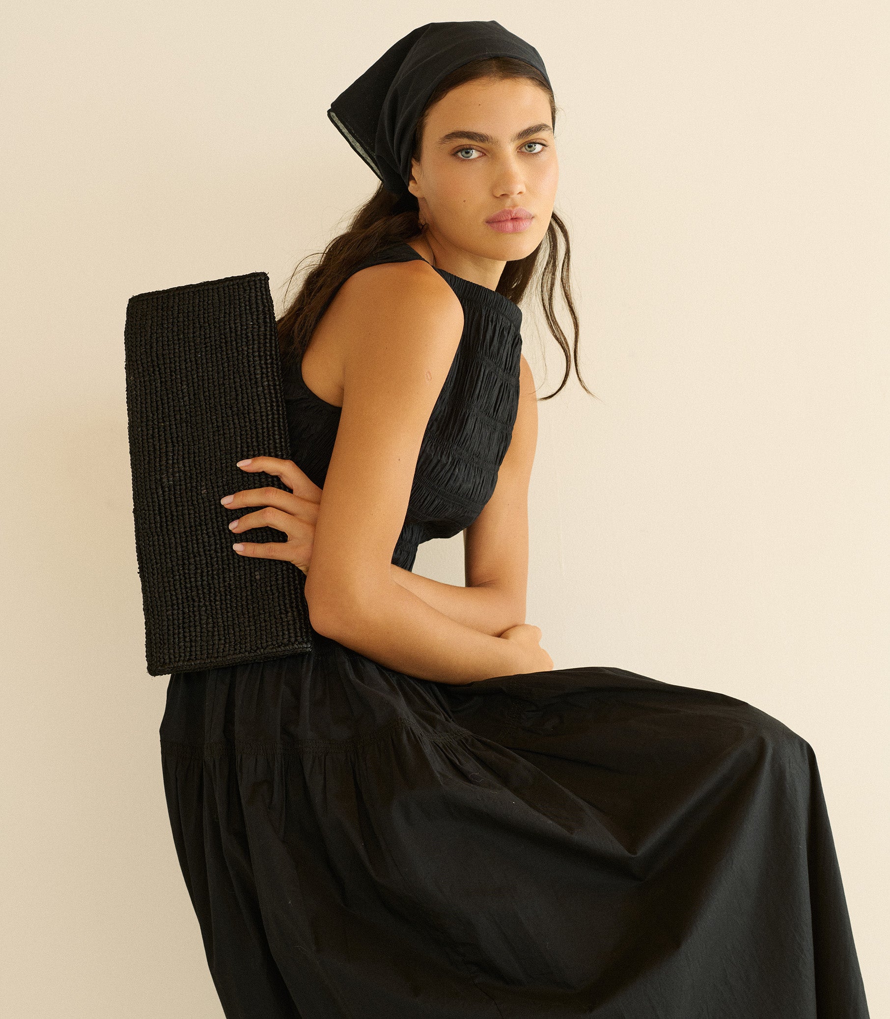 Woman in a black dress holding a black clutch against a beige background